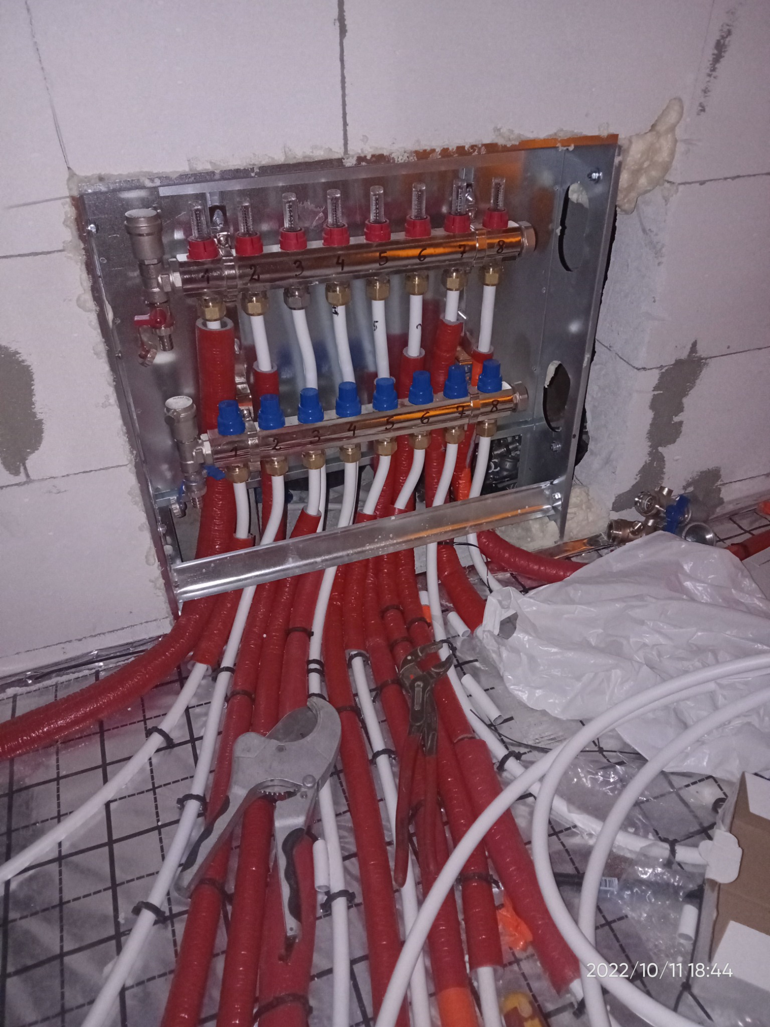 Rozdzielacz ogrzewania podłogowego w trakcie montażu, rury w izolacji czerwonej i białej, widoczne narzędzia do cięcia rur, zamocowany w ścianie z bloczków, pianka montażowa wokół obudowy.