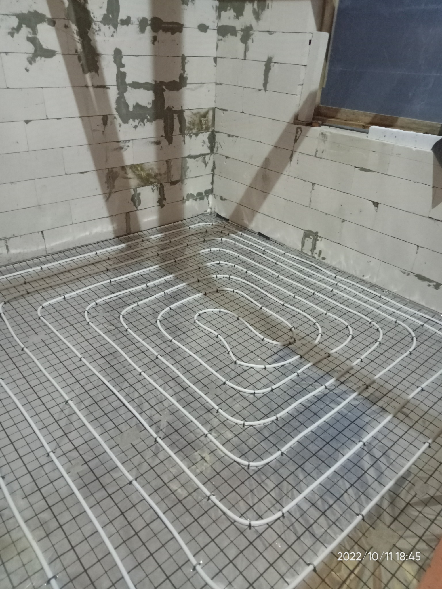 Białe rurki ogrzewania podłogowego ułożone na siatce zbrojeniowej w surowym pomieszczeniu z białych bloczków betonowych.