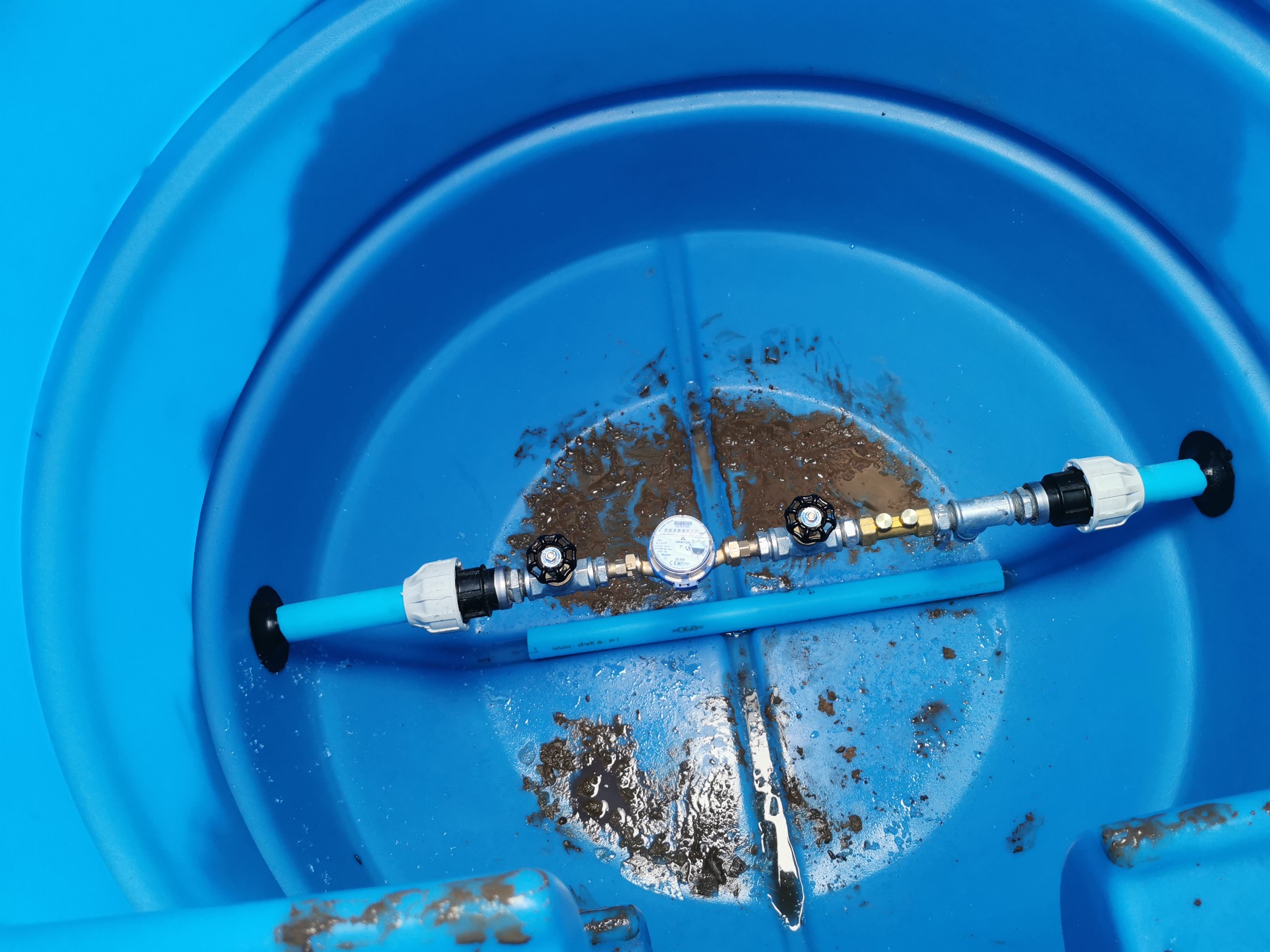 Wnętrze niebieskiego zbiornika z zamontowanym licznikiem i zaworami na niebieskiej rurze, widoczne ślady ziemi i wody.