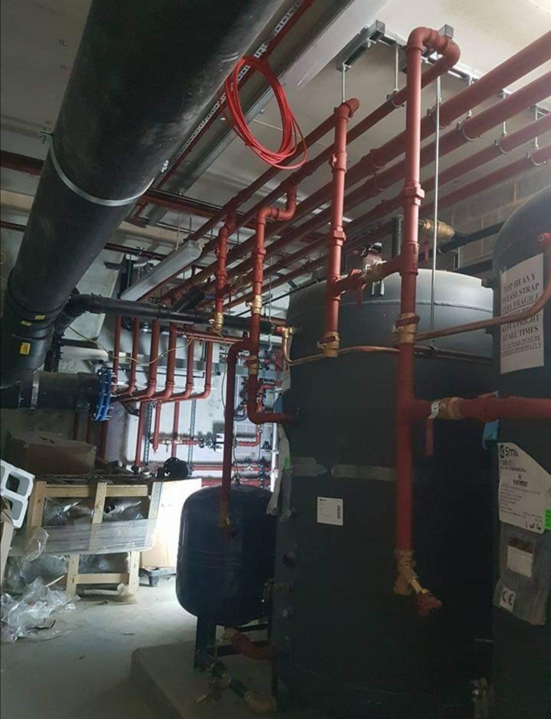 Skomplikowany system rur grzewczych w kolorze czerwonym i czarnym, zainstalowany w pomieszczeniu technicznym z widocznymi zbiornikami i elementami mocującymi do sufitu. Częściowo widoczny bałagan...