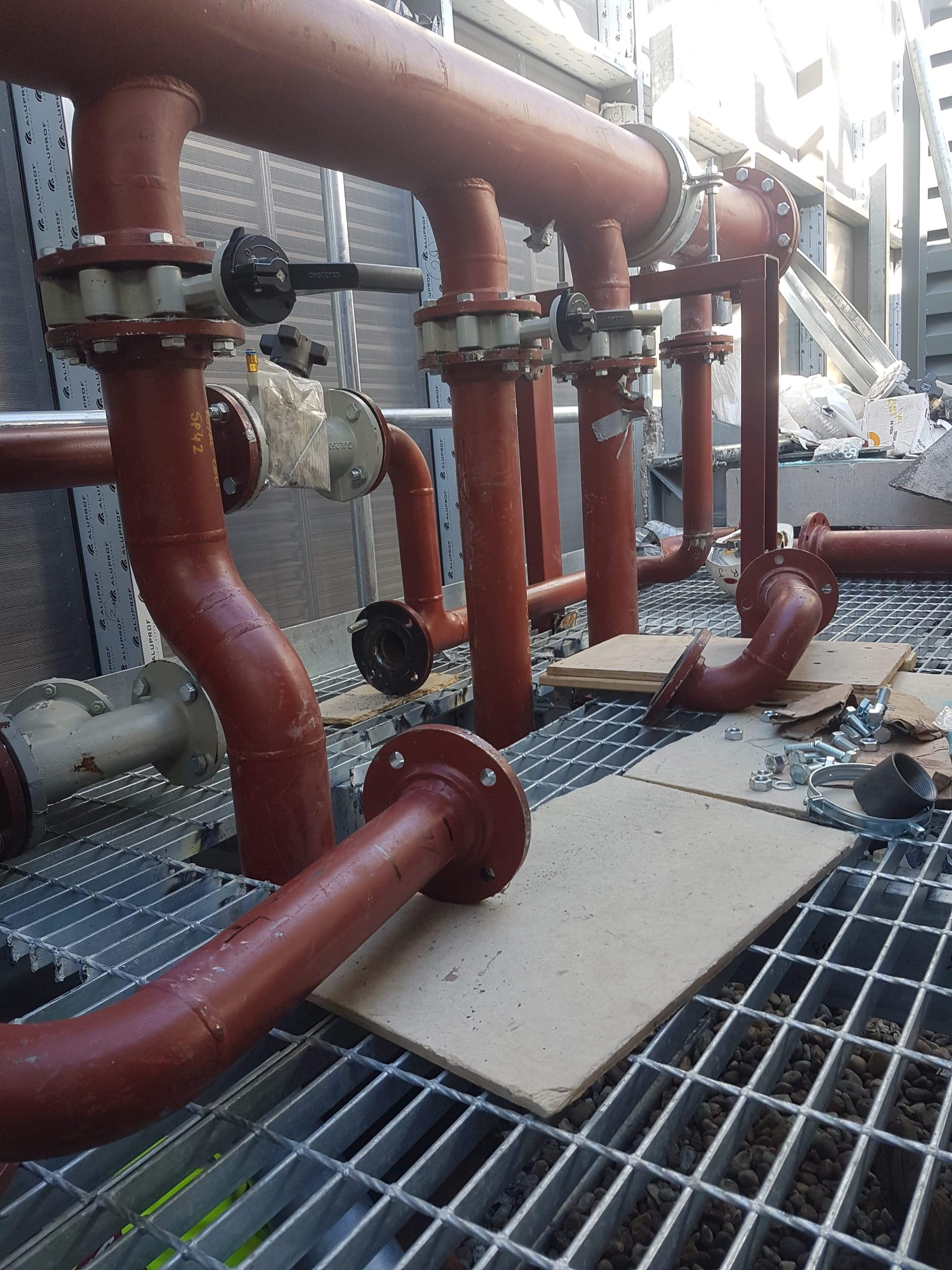 Czerwone rury instalacyjne z zaworami na metalowej kratownicy podłogowej w trakcie montażu, widoczne złącza kołnierzowe, śruby i narzędzia.