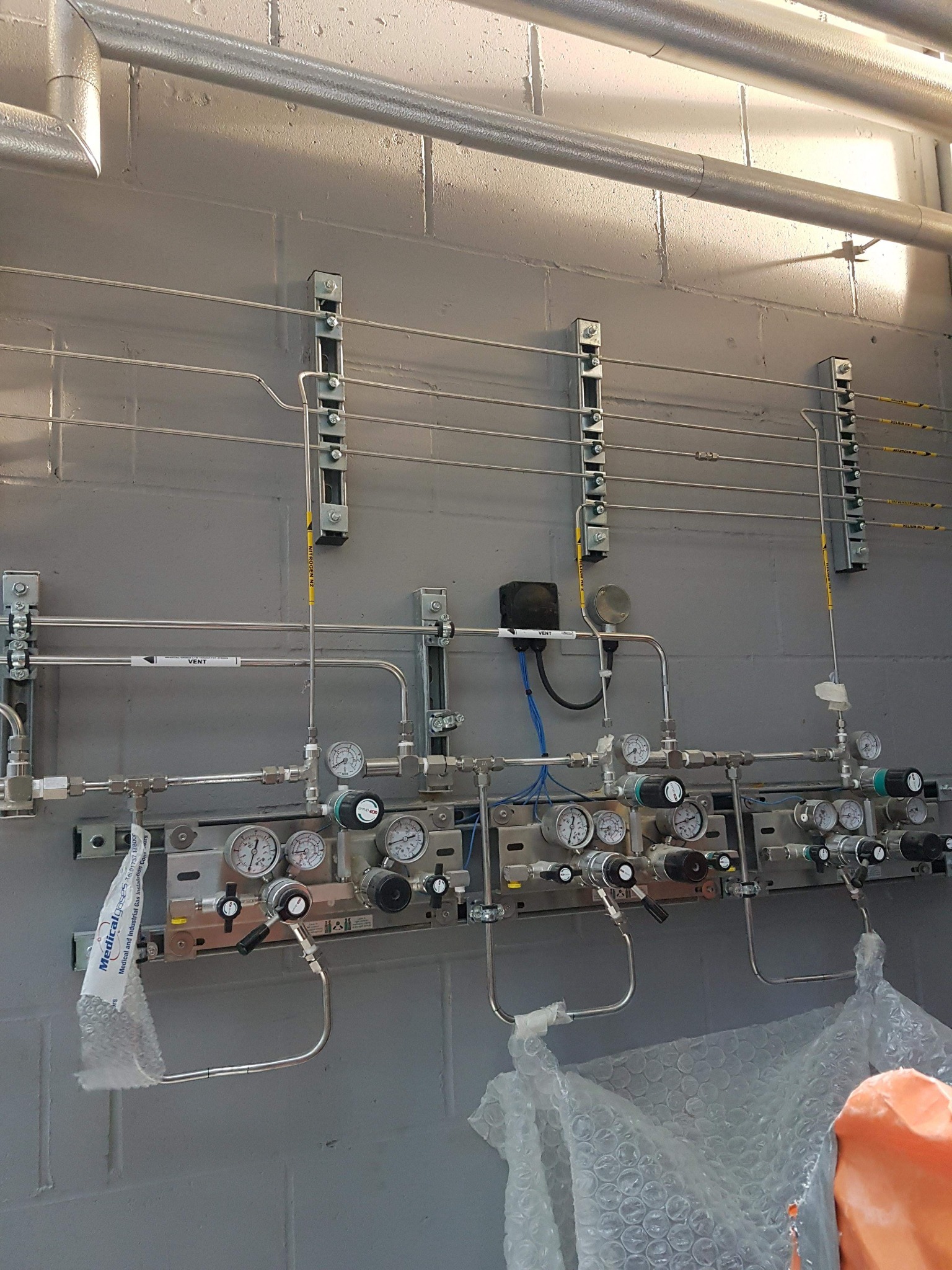 Kompleksowa instalacja gazowa z rurami ze stali nierdzewnej, manometrami i zaworami zamontowana na szarej ścianie z pustaków, z widocznymi oznaczeniami gazów i elementami zabezpieczającymi.