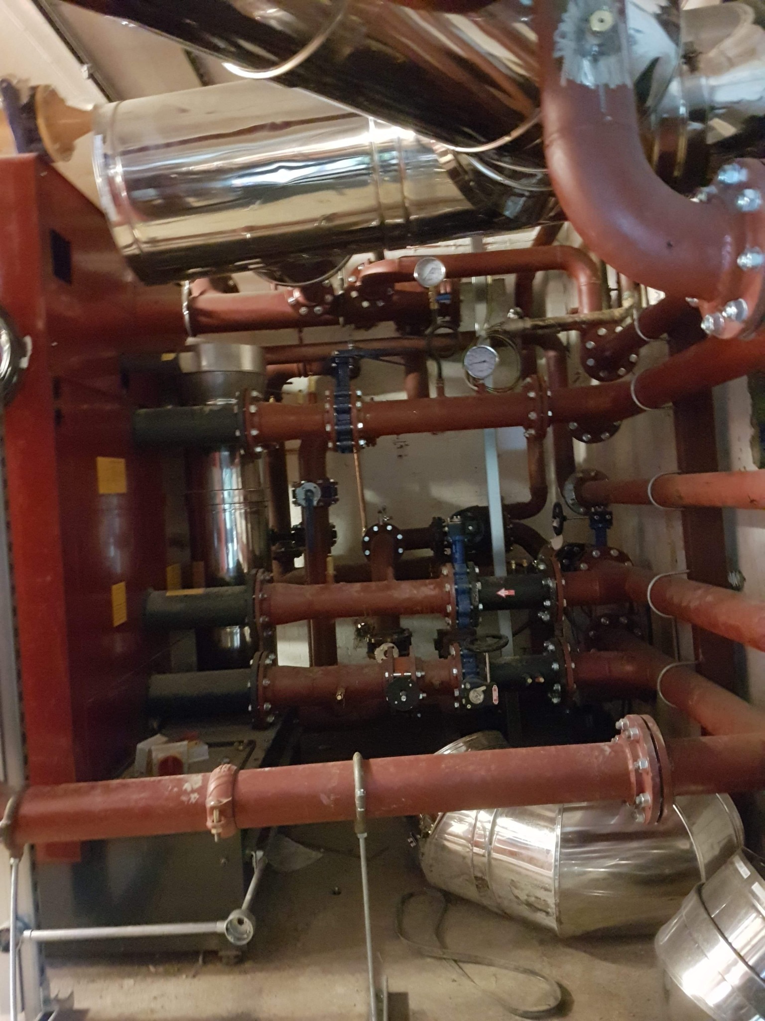 Złożona instalacja rur w kolorze czerwonym i srebrnym z zaworami, manometrami i kołnierzami, widok z góry na skomplikowany system w pomieszczeniu technicznym.