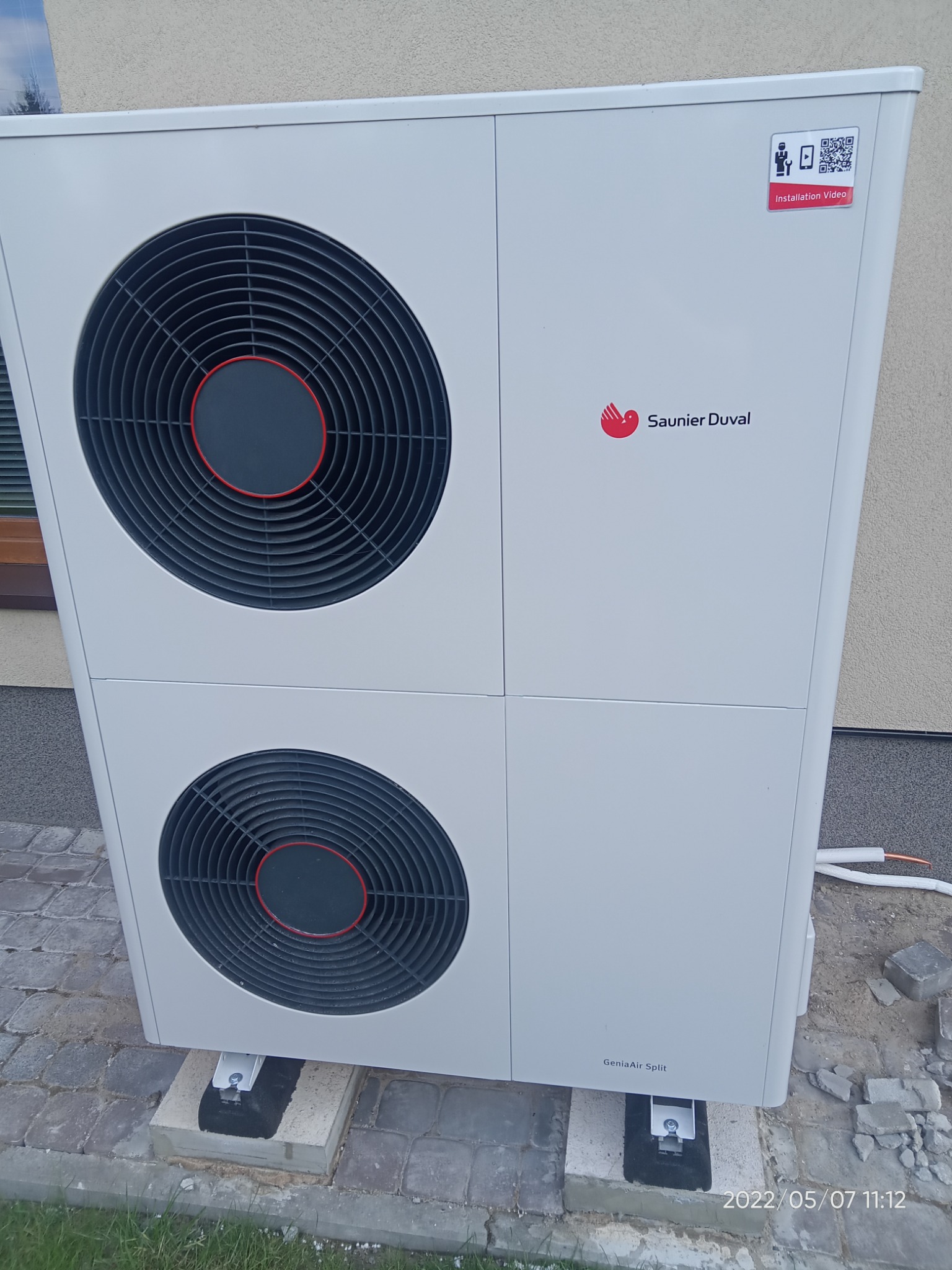 Biała pompa ciepła Saunier Duval GeniaAir Split zamontowana na betonowych podstawach na kostce brukowej, widoczny fragment ściany budynku i przewody instalacyjne.