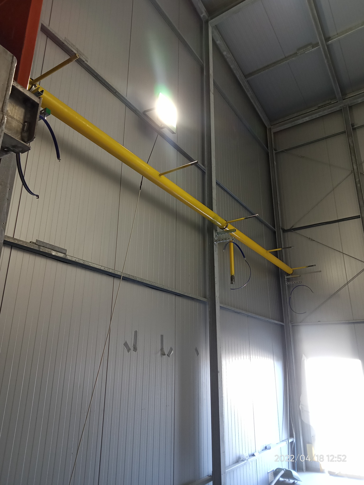 Żółta rura instalacji przemysłowej podwieszona pod sufitem hali z metalowymi ścianami, widoczne elementy mocujące i przewody.