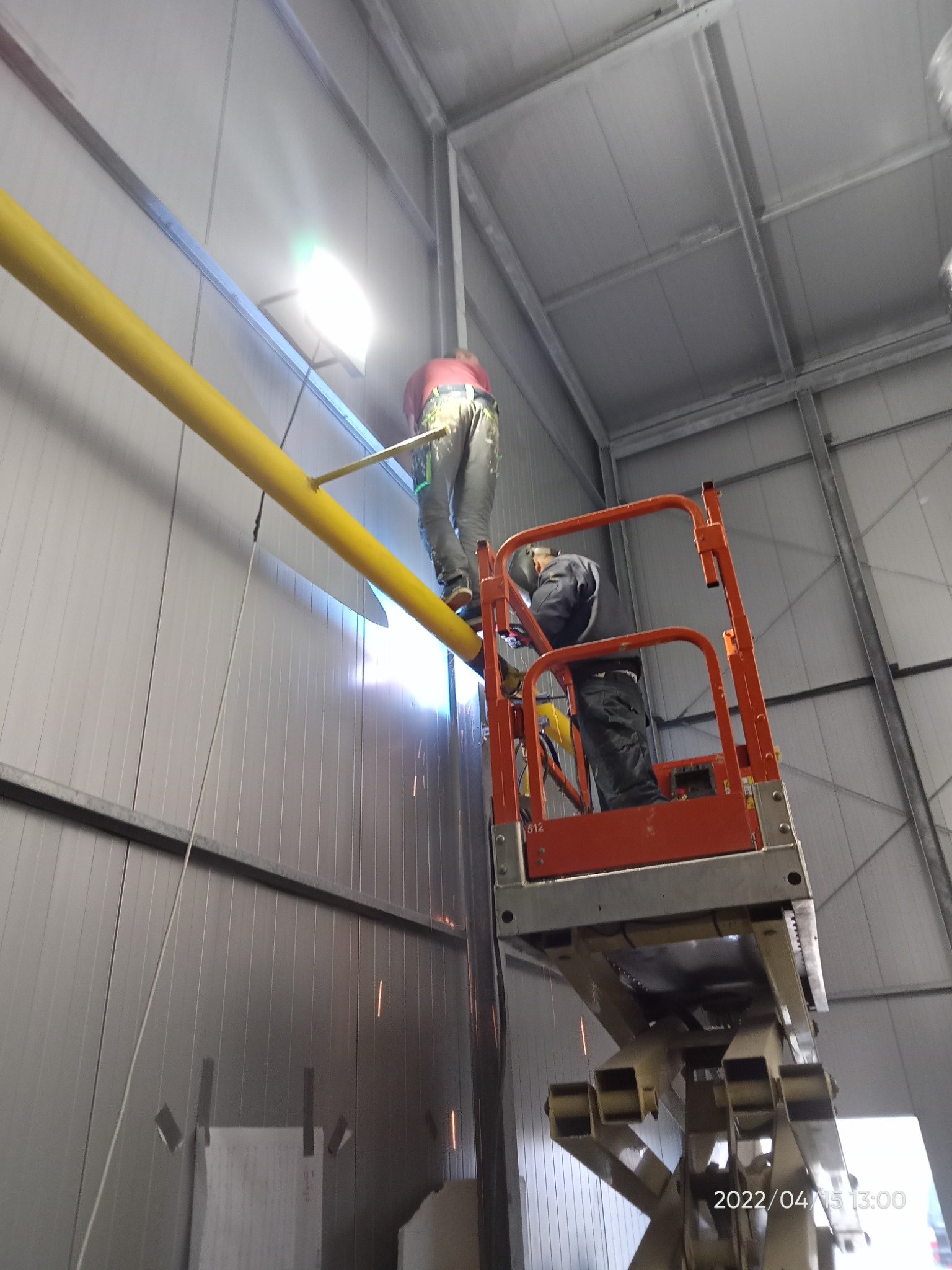 Spawanie żółtej rury gazowej w hali produkcyjnej z podnośnika nożycowego, widoczne iskry i pracownicy w odzieży roboczej.