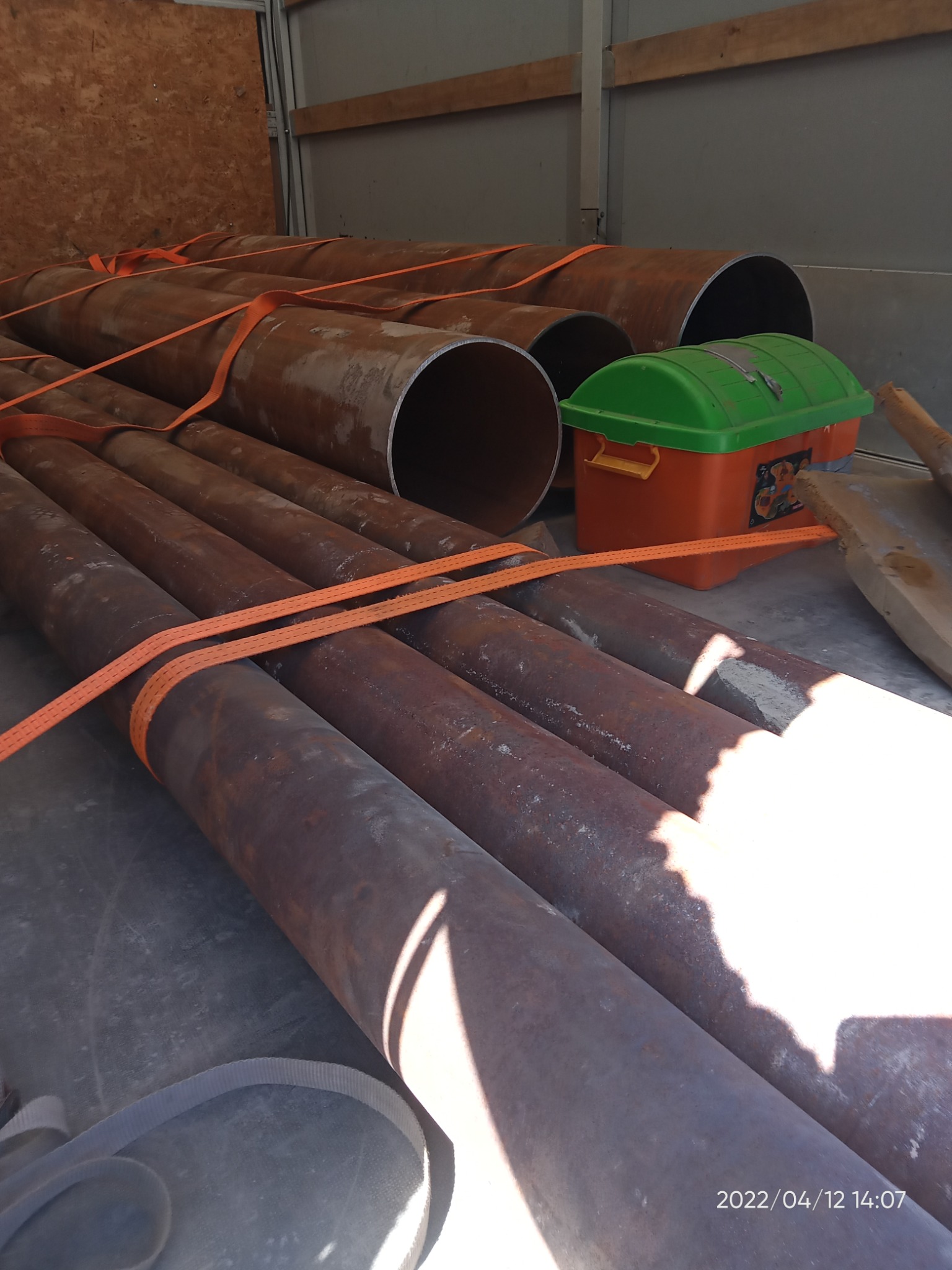 Stalowe rury o dużej średnicy, zabezpieczone pomarańczowymi pasami transportowymi, leżące w przestrzeni ładunkowej pojazdu obok pomarańczowo-zielonej skrzynki narzędziowej.