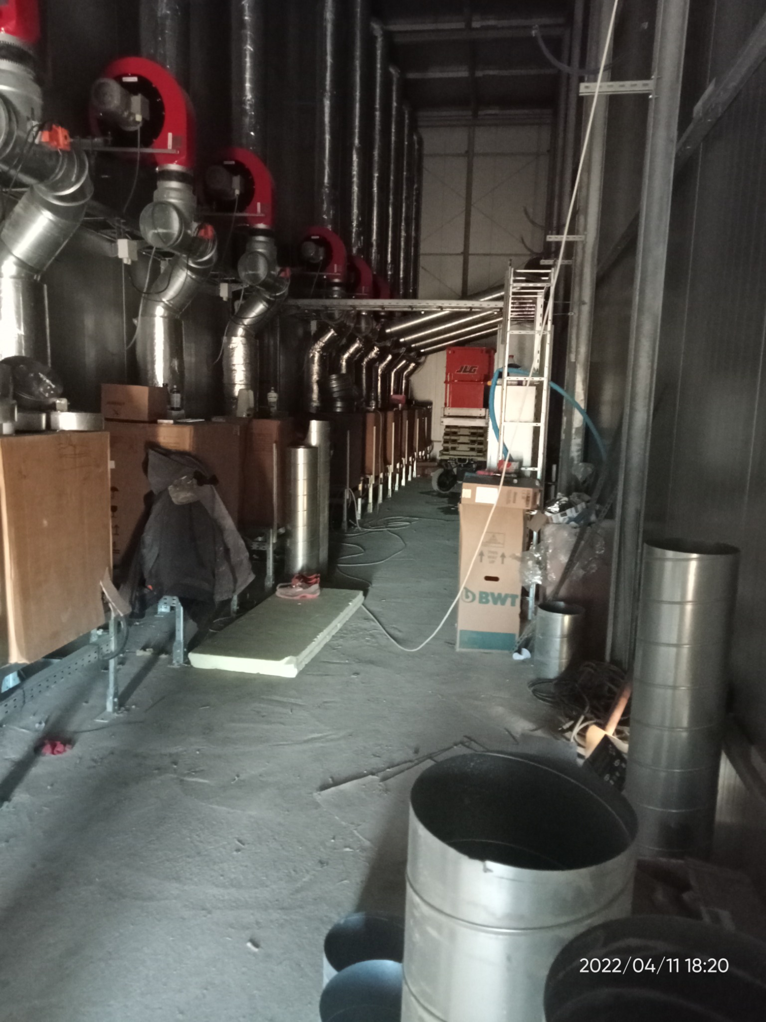 Wnętrze hali z rzędem czerwonych palników i metalowych rur instalacji grzewczej wzdłuż ściany, na podłodze porozrzucane materiały izolacyjne i kartony.