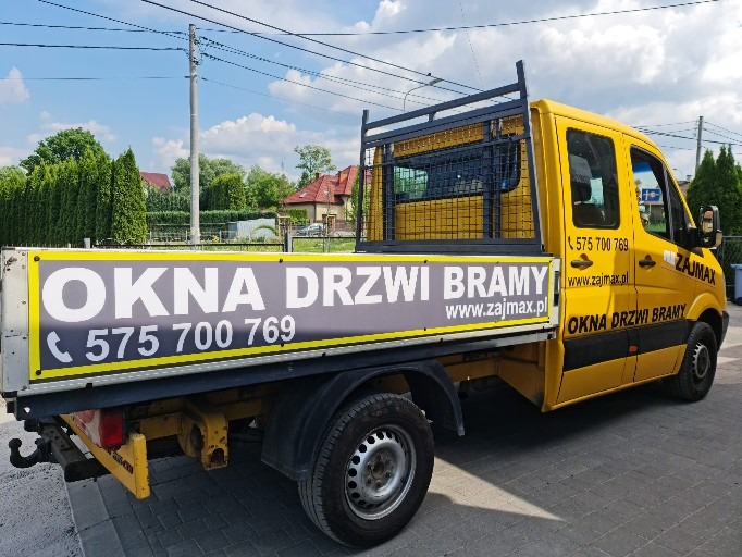 Żółty samochód dostawczy z reklamą okien, drzwi i bram firmy Zajmax, zaparkowany na ulicy w słoneczny dzień.