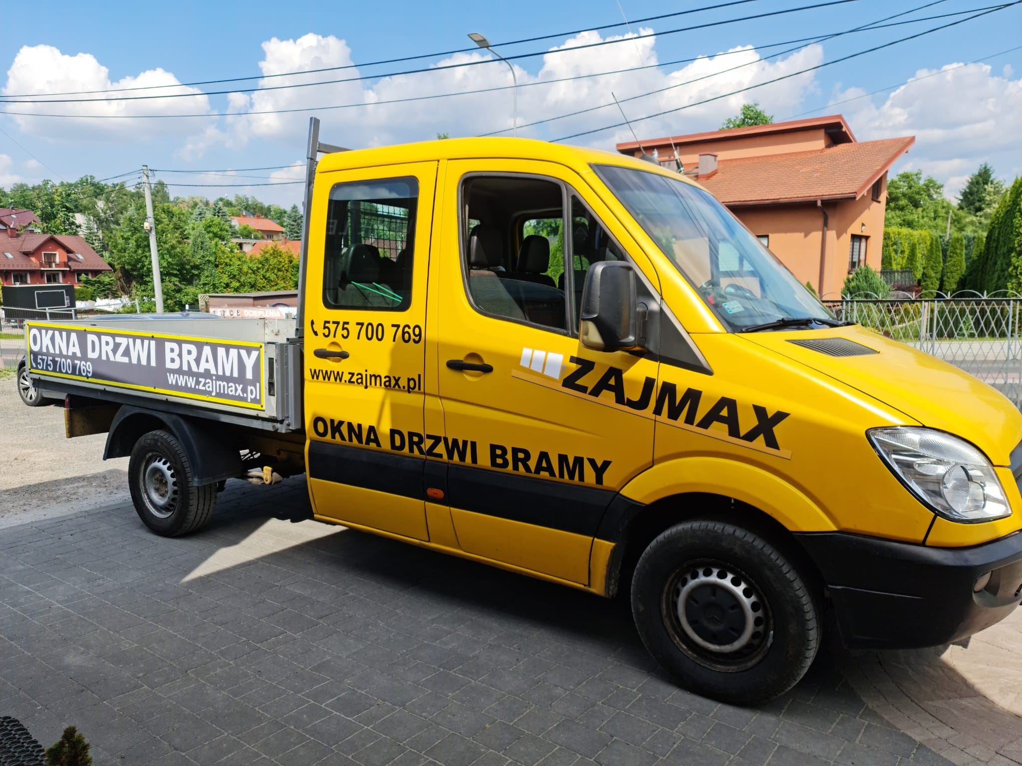 Żółty samochód dostawczy firmy Zajmax z reklamą okien, drzwi i bram na tle domów jednorodzinnych.