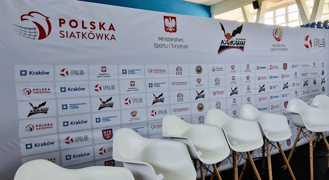 Tło konferencyjne z logotypami, w tym logotypem miasta Kraków, oraz rzędem białych krzeseł barowych. Scena gotowa do konferencji. Grafika stworzona w Krakowie.