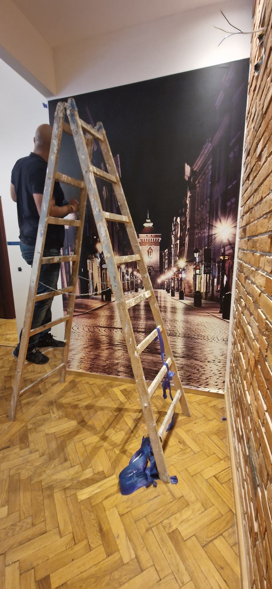 Mężczyzna na drabinie montuje fototapetę z nocnym widokiem miasta na ścianie w pokoju z parkietem i ceglaną ścianą.