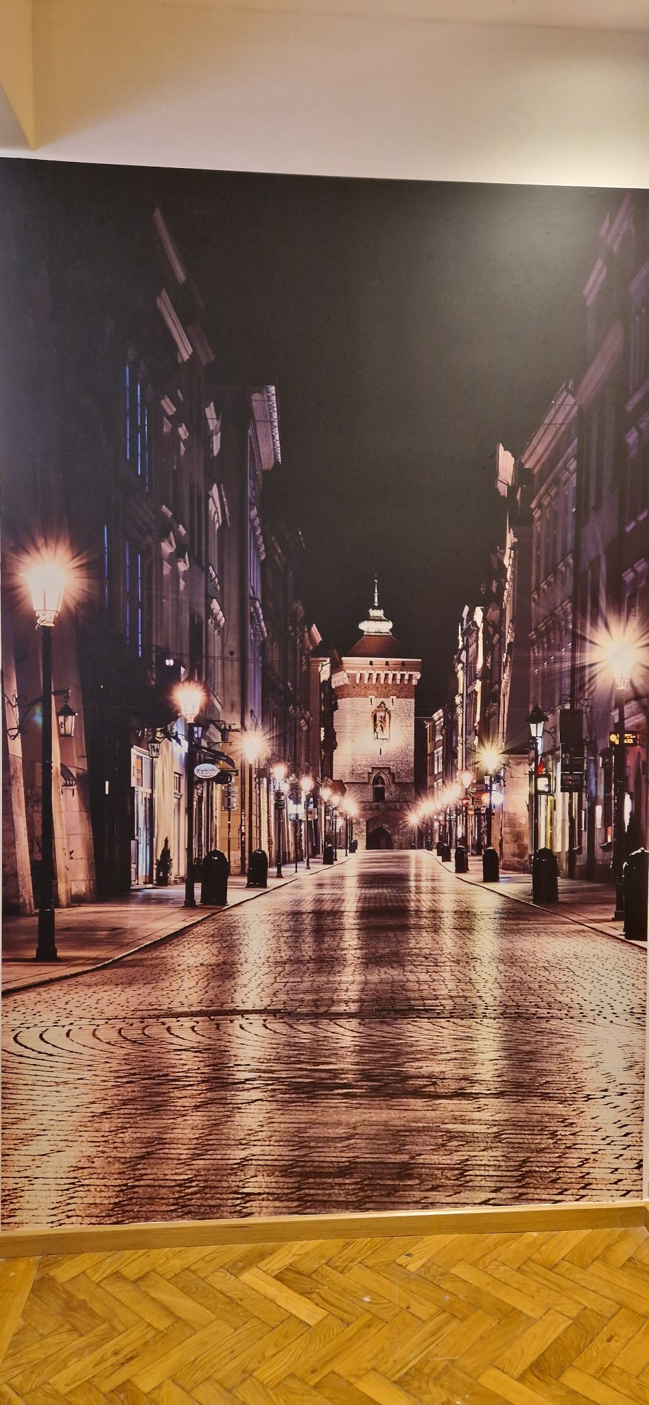 Fototapeta przedstawiająca nocny widok na ulicę z brukowaną nawierzchnią i wieżą w oddali, oświetloną przez latarnie, umieszczona na ścianie nad parkietem.