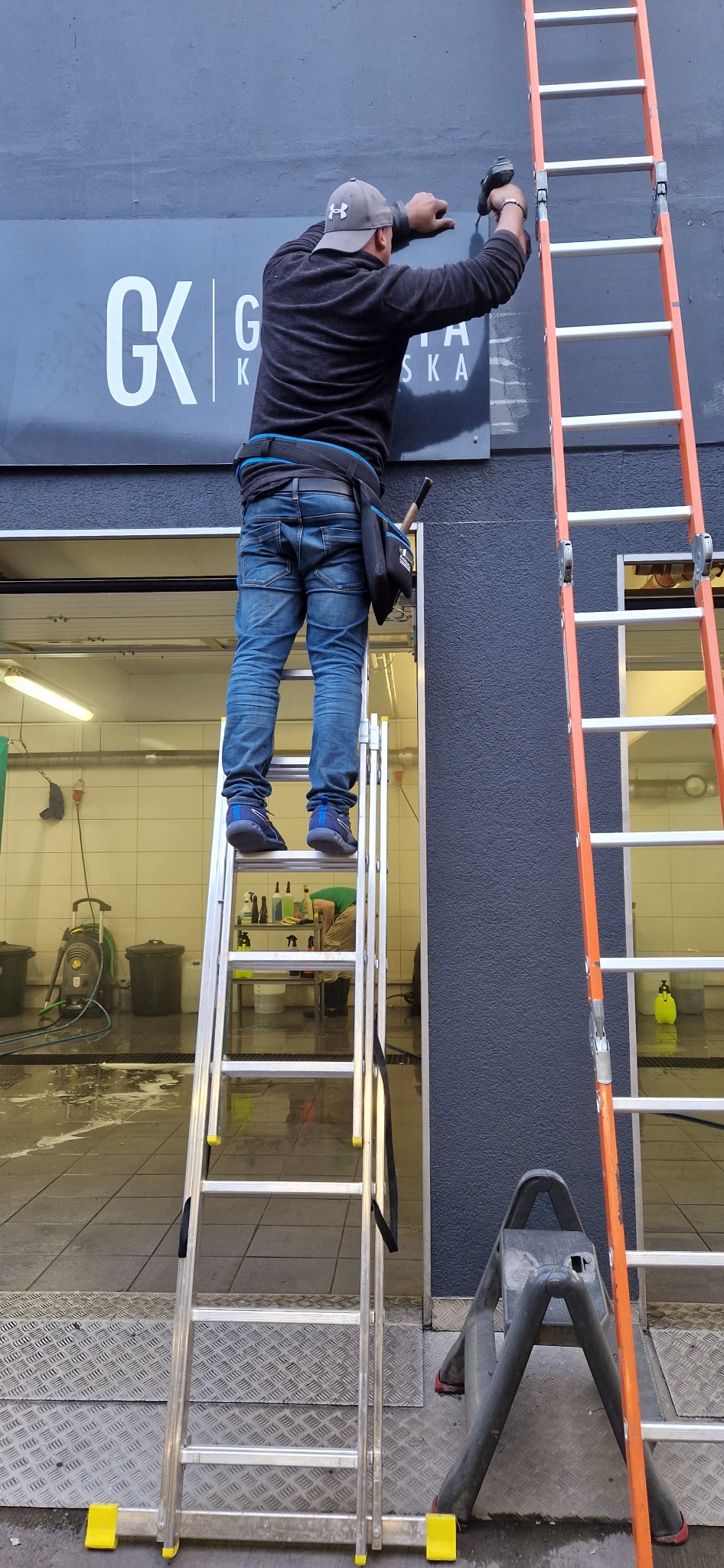 Mężczyzna na drabinie montuje szyld z napisem 'CK' na szarej elewacji budynku, widoczna druga drabina i wnętrze myjni samochodowej.