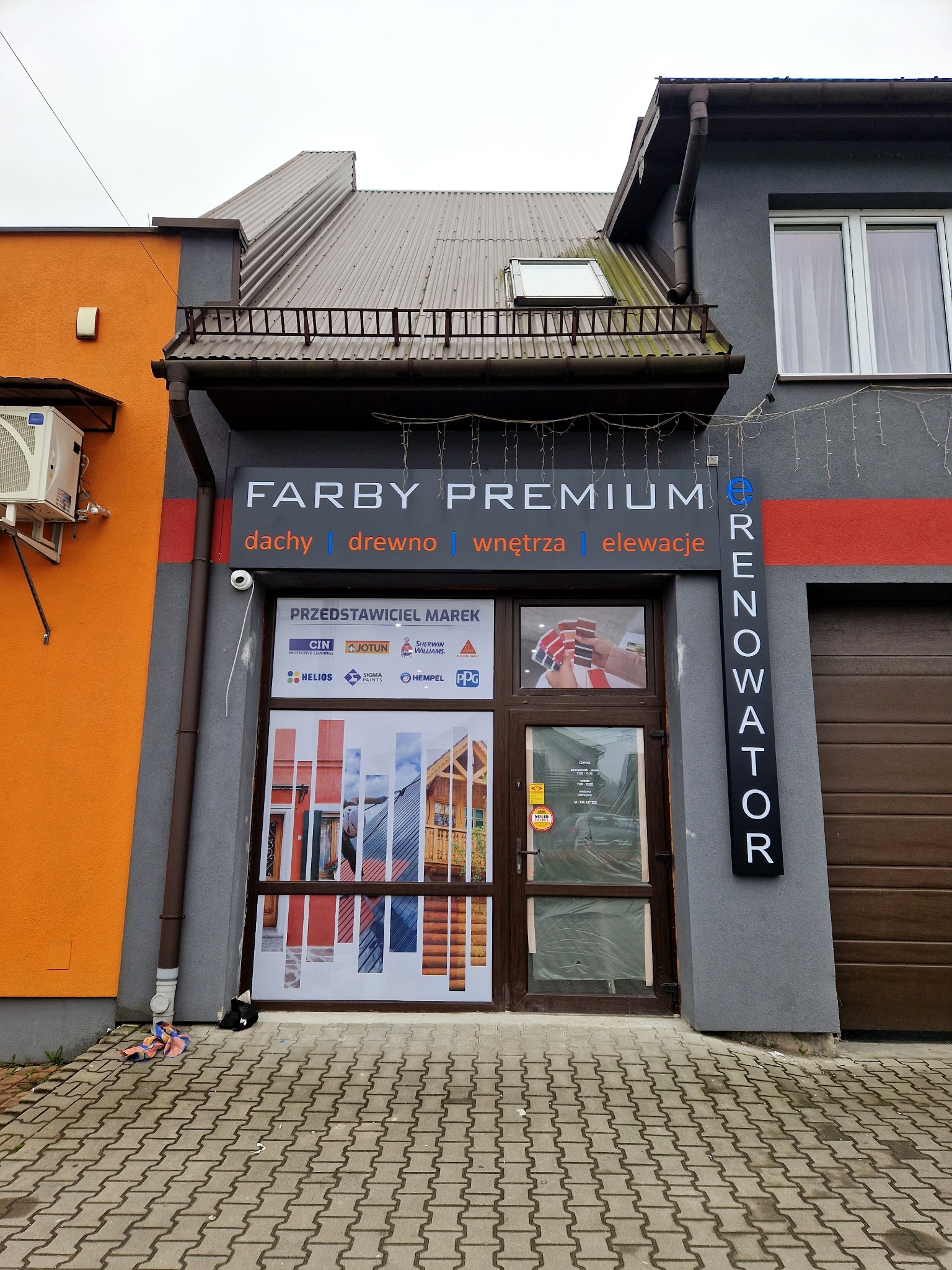 Fasada sklepu z farbami 'Farby Premium', prezentująca materiały do dachów, drewna, wnętrz i elewacji. W oknie naklejki z prezentacją produktów i logo producentów farb. Obok wejścia pionowy szyld...