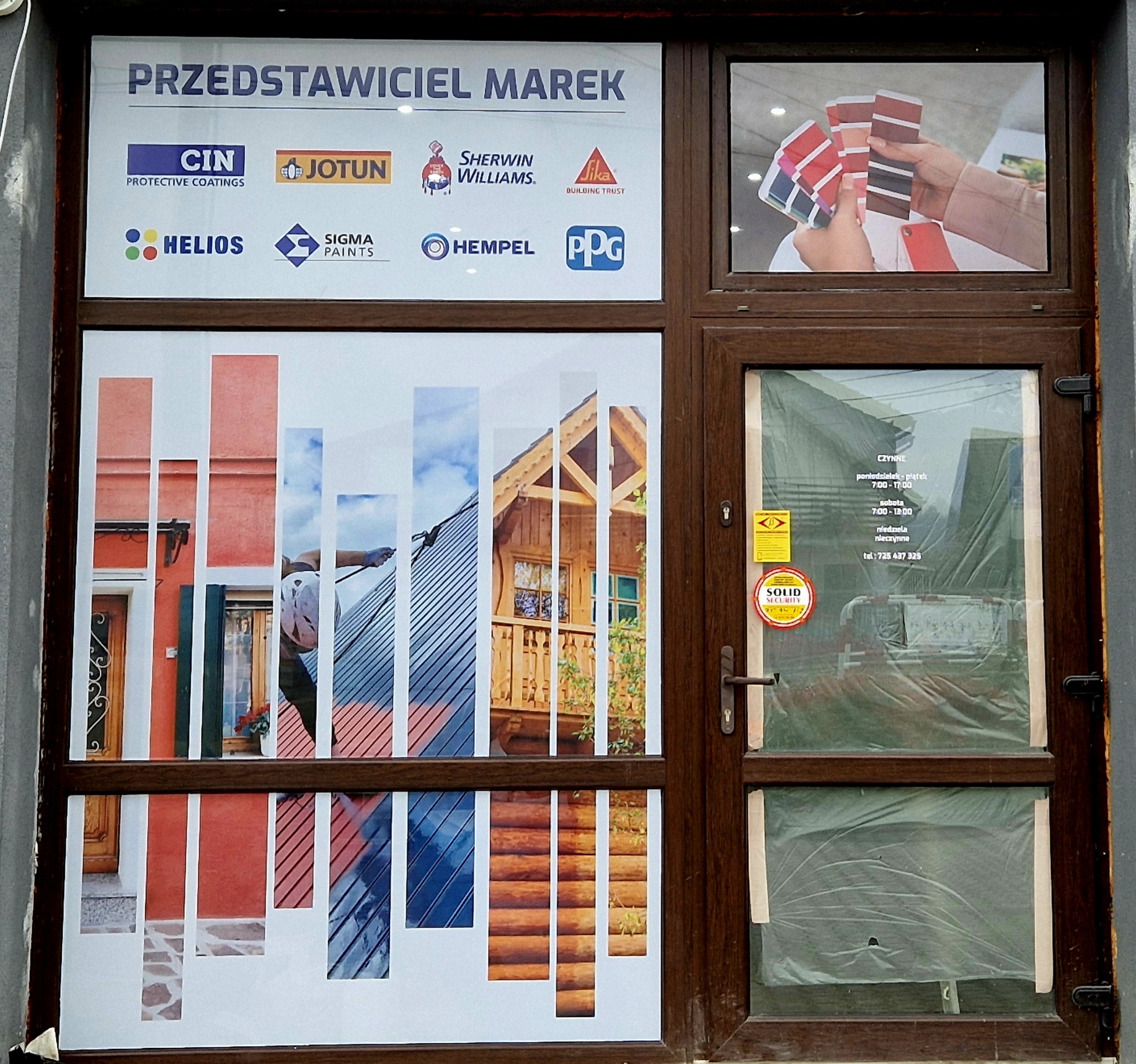 Okno punktu sprzedaży farb z naklejonymi reklamami różnych marek, w tym palety kolorów trzymane w dłoniach