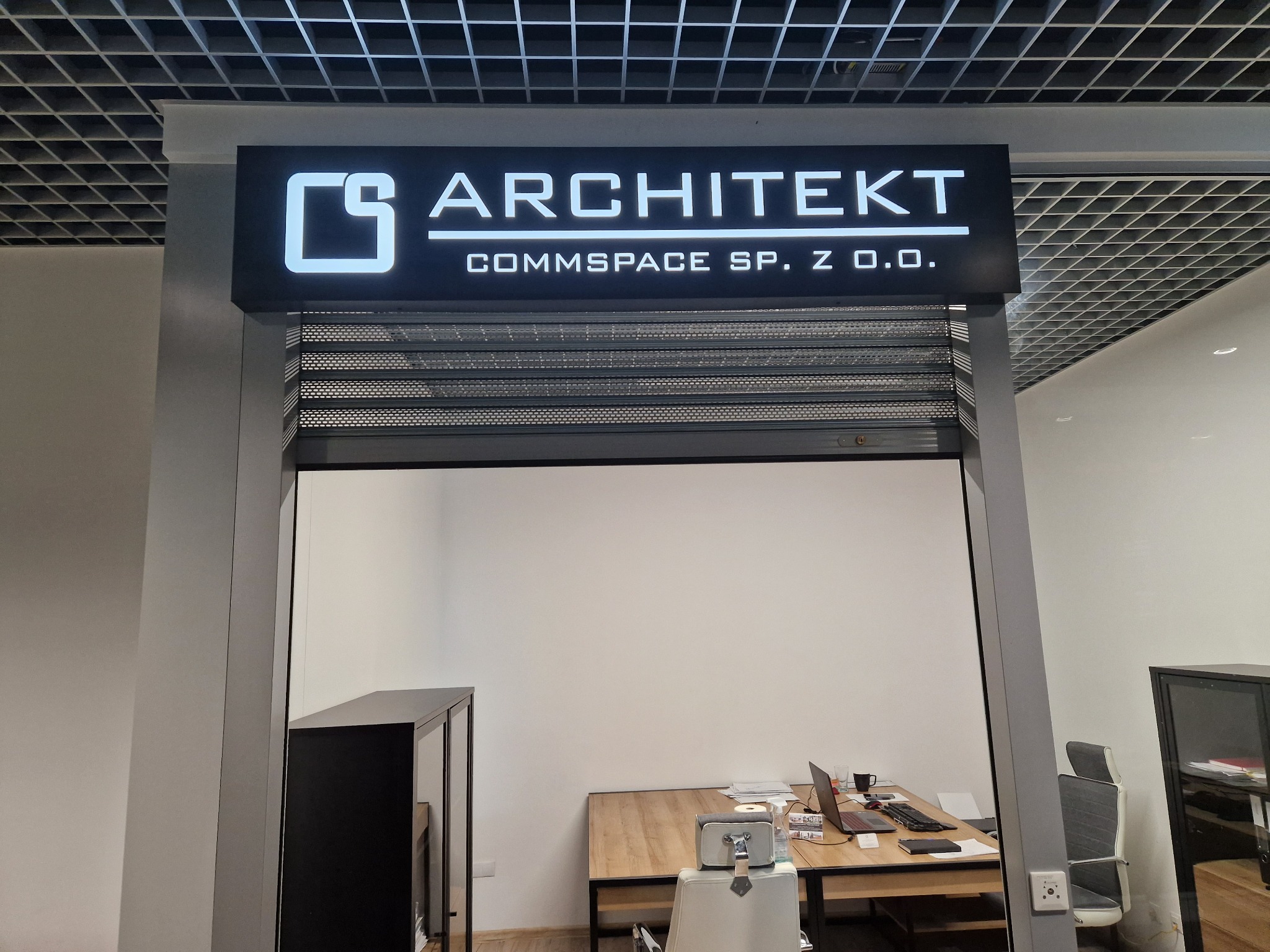 Elegancki szyld świetlny z logo i nazwą firmy ARCHITEKT COMMSPACE SP. Z O.O. nad wejściem do biura z widocznym biurkiem, laptopem i krzesłem w tle.