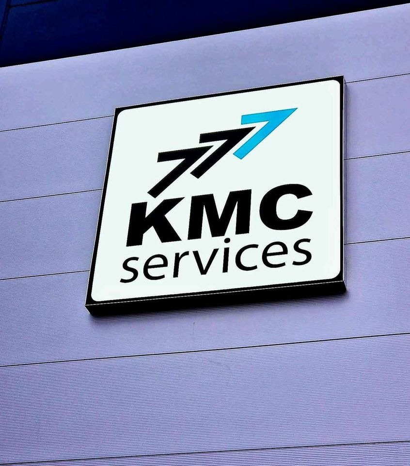 Kwadratowy szyld firmy KMC Services z trzema czarnymi strzałkami i jedną niebieską, zamontowany na ścianie z poziomymi panelami.