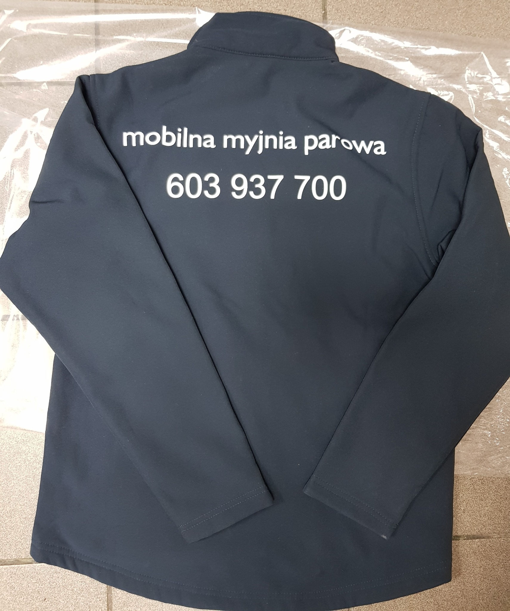Granatowa bluza robocza z długimi rękawami, na plecach biały nadruk 'mobilna myjnia parowa' i numer telefonu 603 937 700, leżąca na podłodze zabezpieczonej folią.