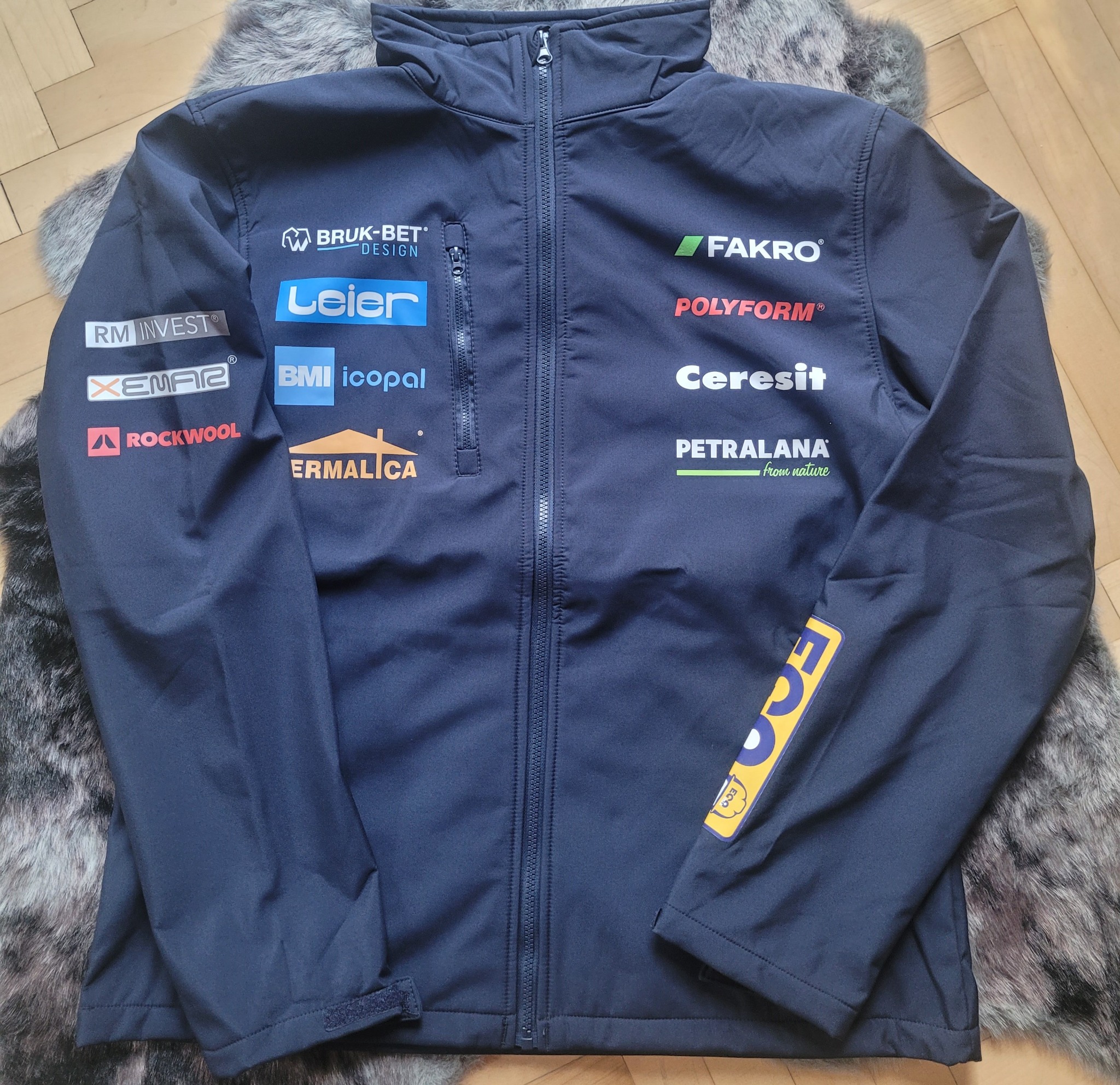 Granatowa kurtka softshell z licznymi nadrukami logo firm budowlanych, leżąca na futrzanym dywaniku.