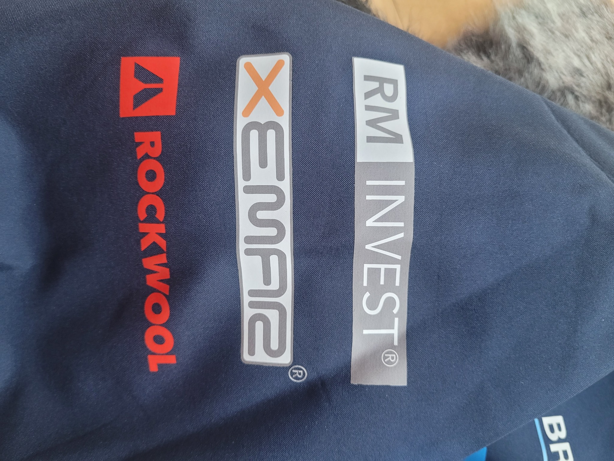Granatowa kurtka robocza z trzema pionowymi nadrukami: czerwonym logo Rockwool, szarym Xemar z pomarańczowym X oraz szarym RM Invest.