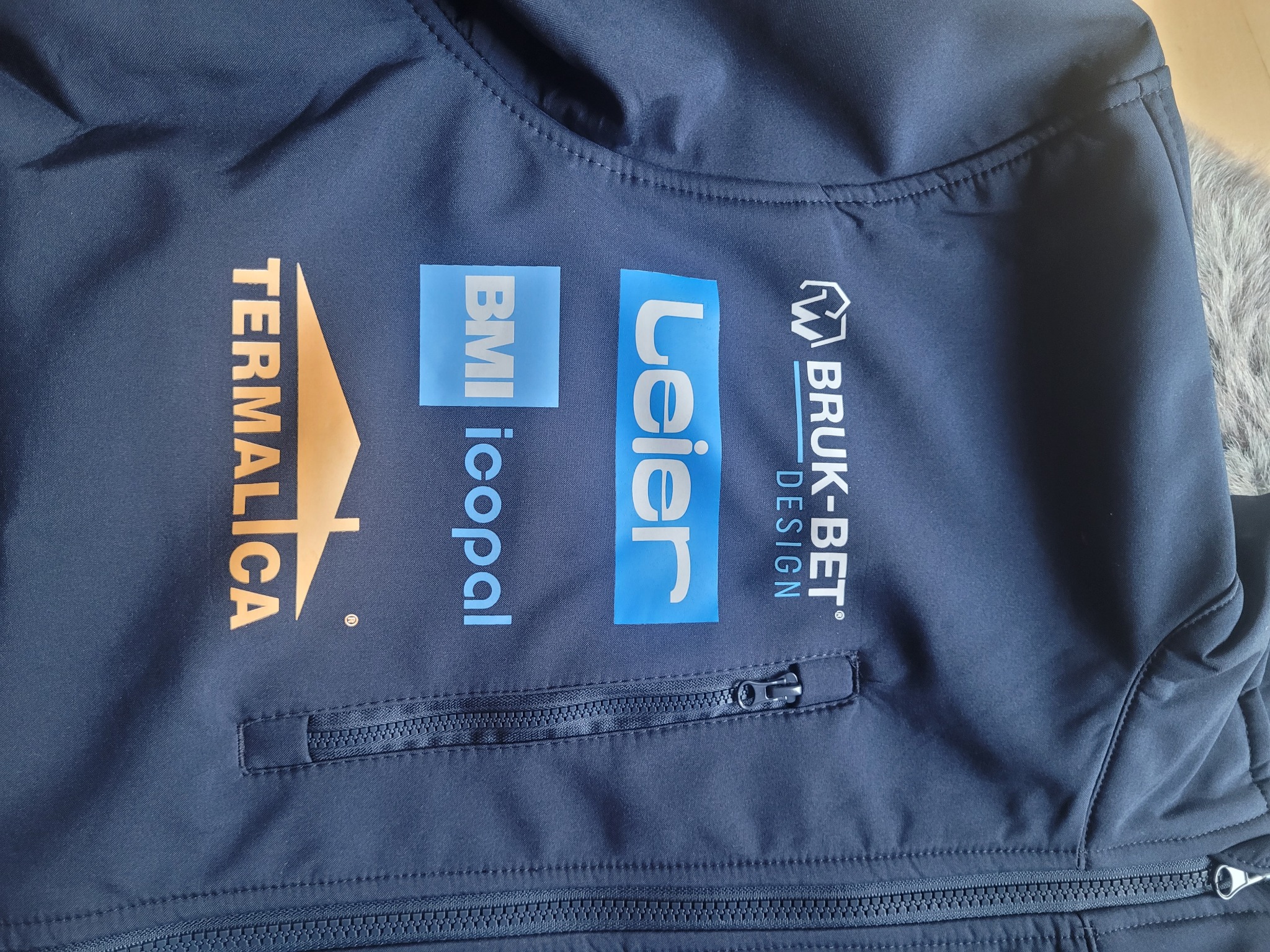 Granatowa kurtka softshell z nadrukowanymi logotypami firm: Termalica, BMI Icopal, Leler i Bruk-Bet Design, widoczny zamek błyskawiczny kieszeni.
