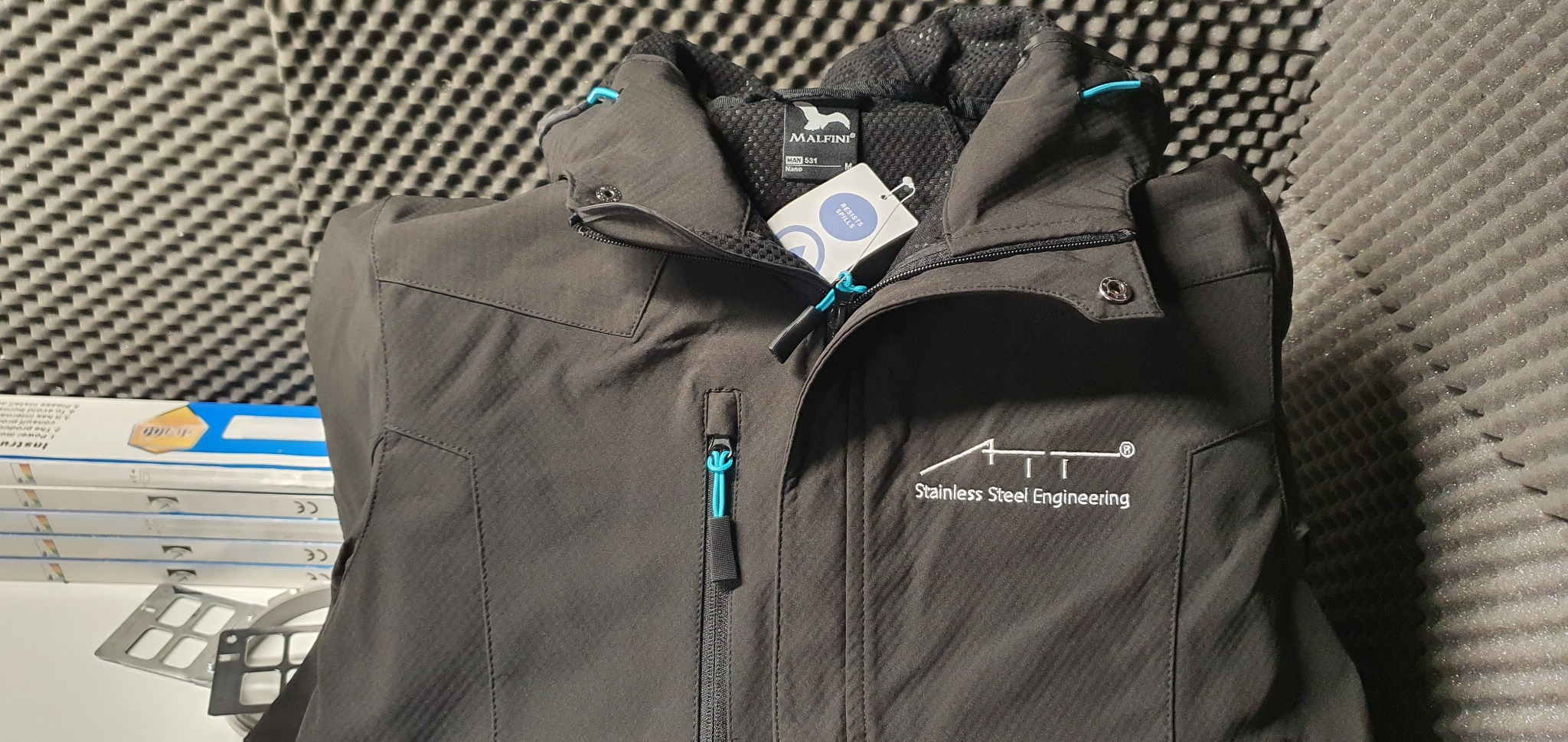 Czarna kurtka robocza marki Malfini z nadrukowanym logo 'Stainless Steel Engineering', widoczny zamek błyskawiczny z turkusowym akcentem, tło z pianki akustycznej.