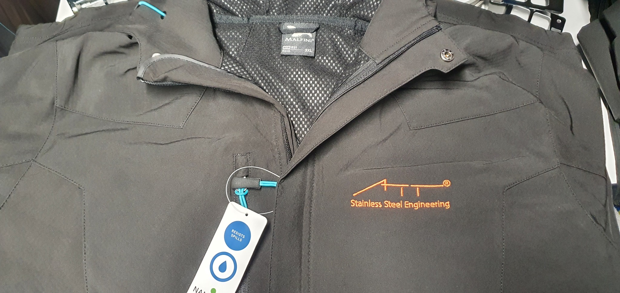 Czarna kurtka robocza z pomarańczowym logo 'Stainless Steel Engineering', widoczna metka z informacją o odporności na plamy, zamek błyskawiczny i siateczkowa podszewka wewnątrz.