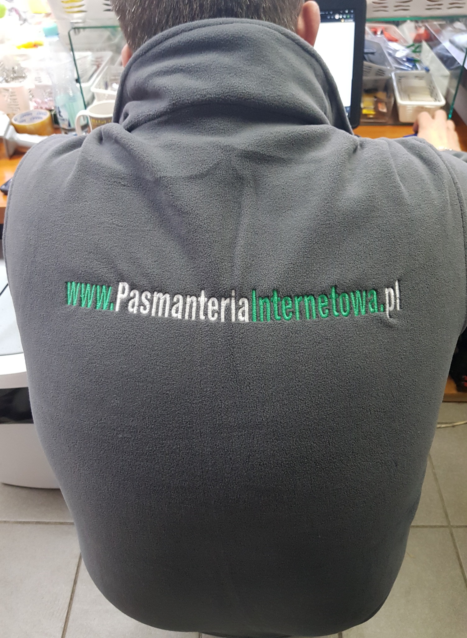 Tył szarej polarowej kamizelki z wyhaftowanym adresem strony internetowej www.PasmanteriaInternetowa.pl, widoczne ramiona osoby siedzącej przy biurku z komputerem.