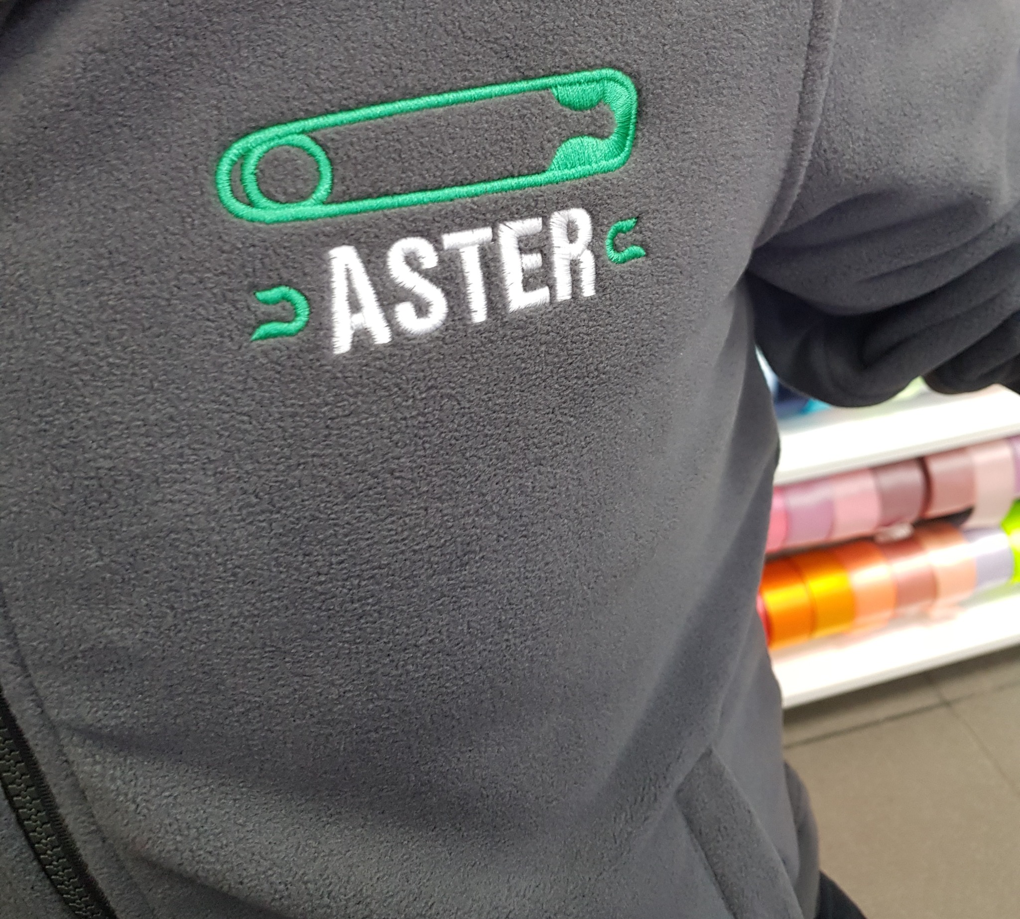 Szara bluza polarowa z haftowanym logo firmy 'ASTER' w postaci agrafki i nazwy, widoczne z bliska detale ściegów.
