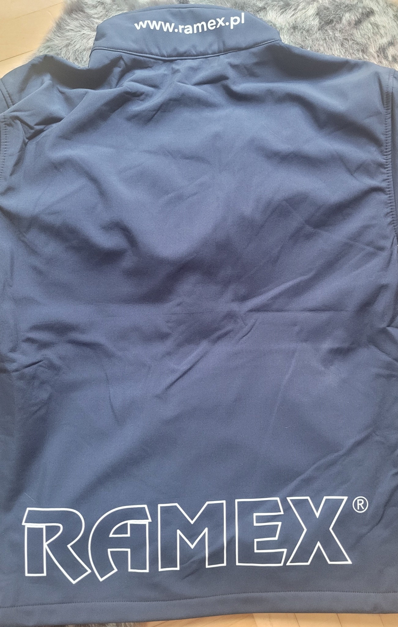Granatowa kurtka robocza z białym napisem 'RAMEX' i adresem strony internetowej na karku, leżąca na futrzanym tle.