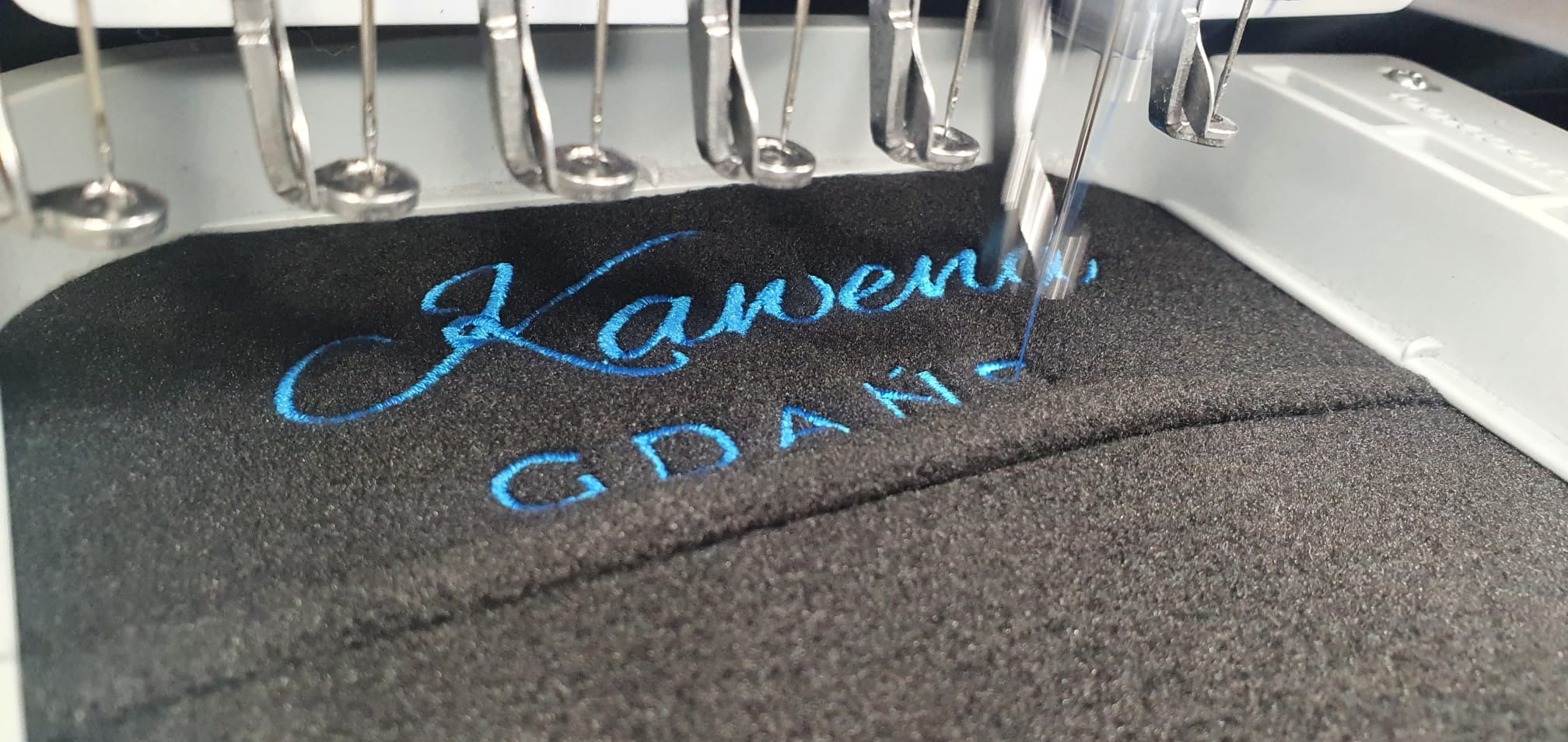 Proces haftowania niebieskiego napisu 'Kawena GDAŃSK' na czarnym, szorstkim materiale za pomocą przemysłowej maszyny do haftowania.