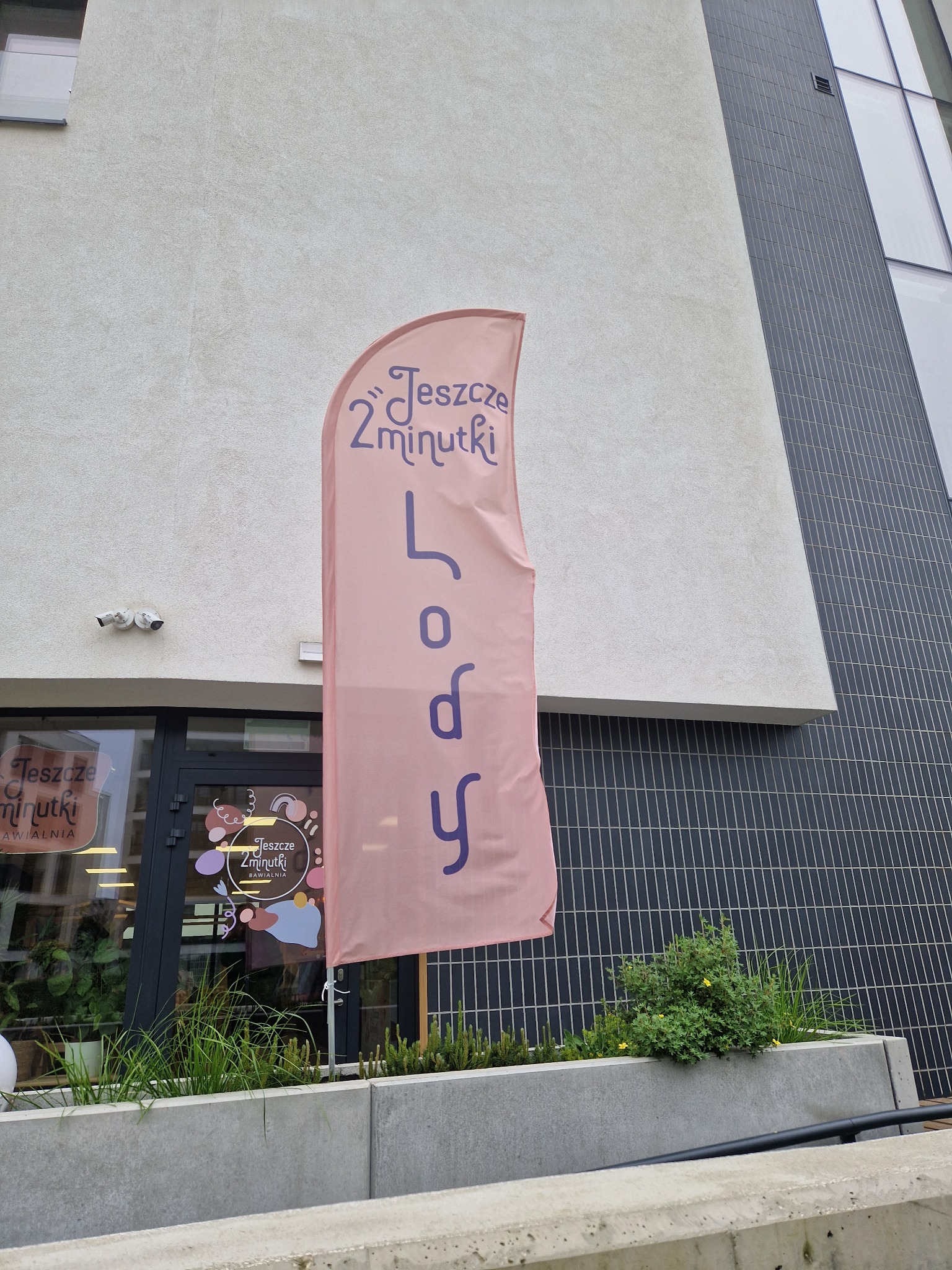 Pionowy, różowy baner reklamowy z napisem 'Jeszcze 2 minutki Lody' przed wejściem do lokalu, widok z dołu, nowoczesna architektura w tle.