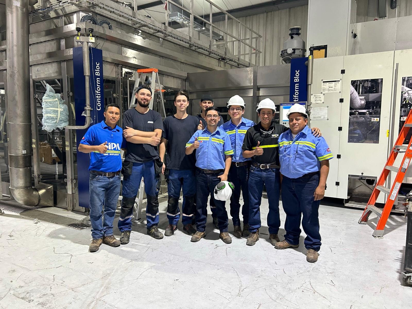 Grupa ośmiu mężczyzn w odzieży roboczej i kaskach, stojących przed przemysłową maszyną Contiform Bloc, z drabiną w tle, wykonujących gest kciuka w górę.