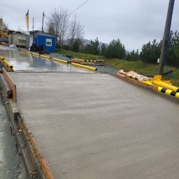 BORYSBUD Natan Borysiewicz - Świeżo wylany, mokry beton w szalunku, widok z góry. W tle budka, drzewa i pochmurne niebo. Żółte elementy zabezpieczające na krawędziach.