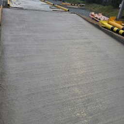 BORYSBUD Natan Borysiewicz - Świeżo wylany, gładki betonowy fundament z żółtymi rurami ochronnymi na obrzeżach, w tle budynek i zieleń. Widok z góry, pochmurne niebo.