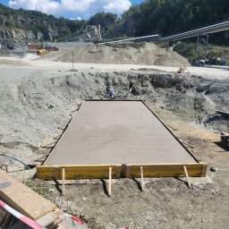 BORYSBUD Natan Borysiewicz - Wylany fundament w szalunku na tle kamieniołomu. Widoczne deski szalunkowe, świeży beton i otoczenie placu budowy z maszynami i hałdami ziemi.