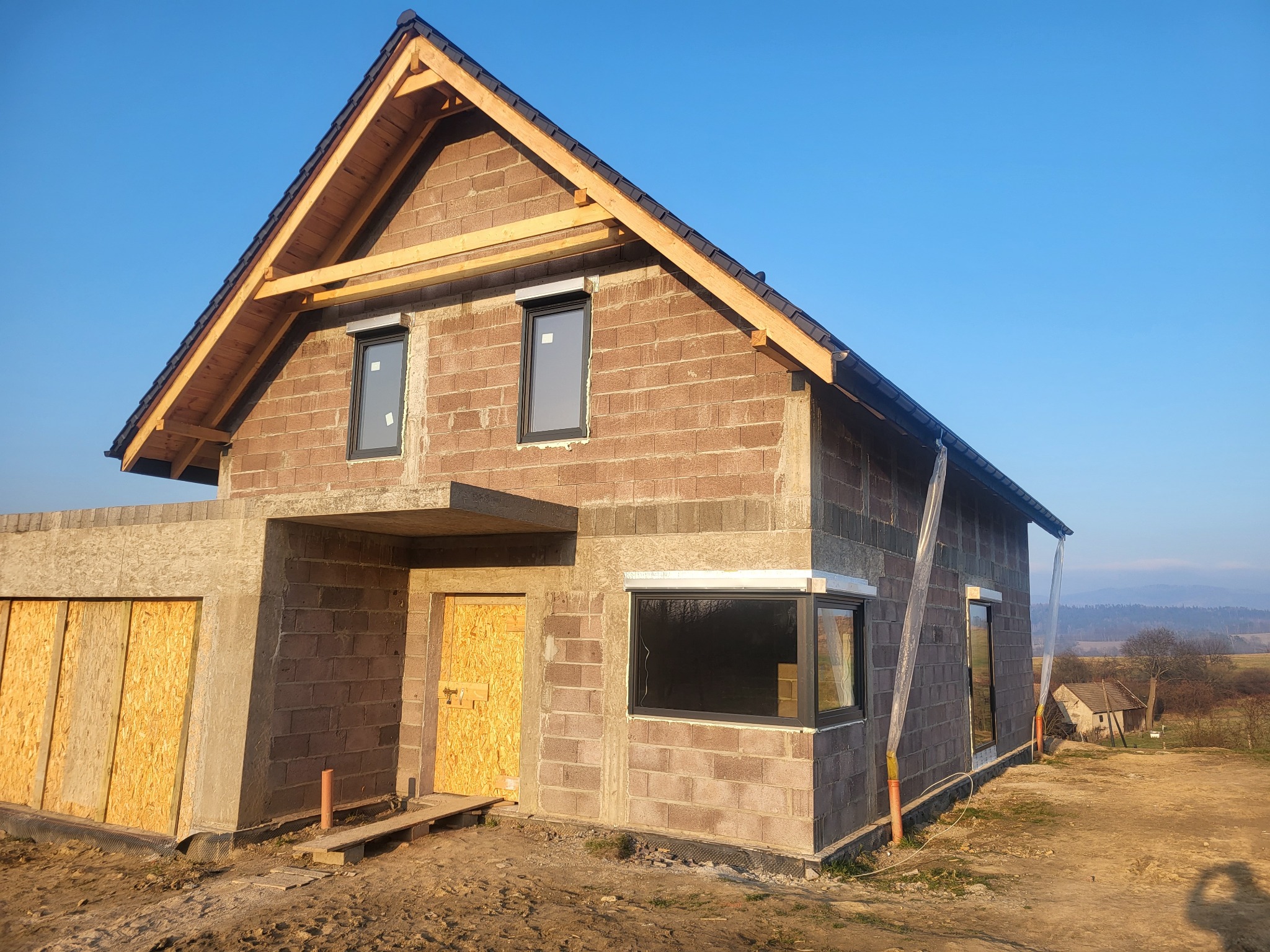 Surowy, dwukondygnacyjny dom z keramzytu w trakcie budowy, z zamontowanymi oknami w ciemnych ramach, zabezpieczonymi otworami drzwiowymi płytami OSB i widoczną konstrukcją dachu, na tle błękitnego...