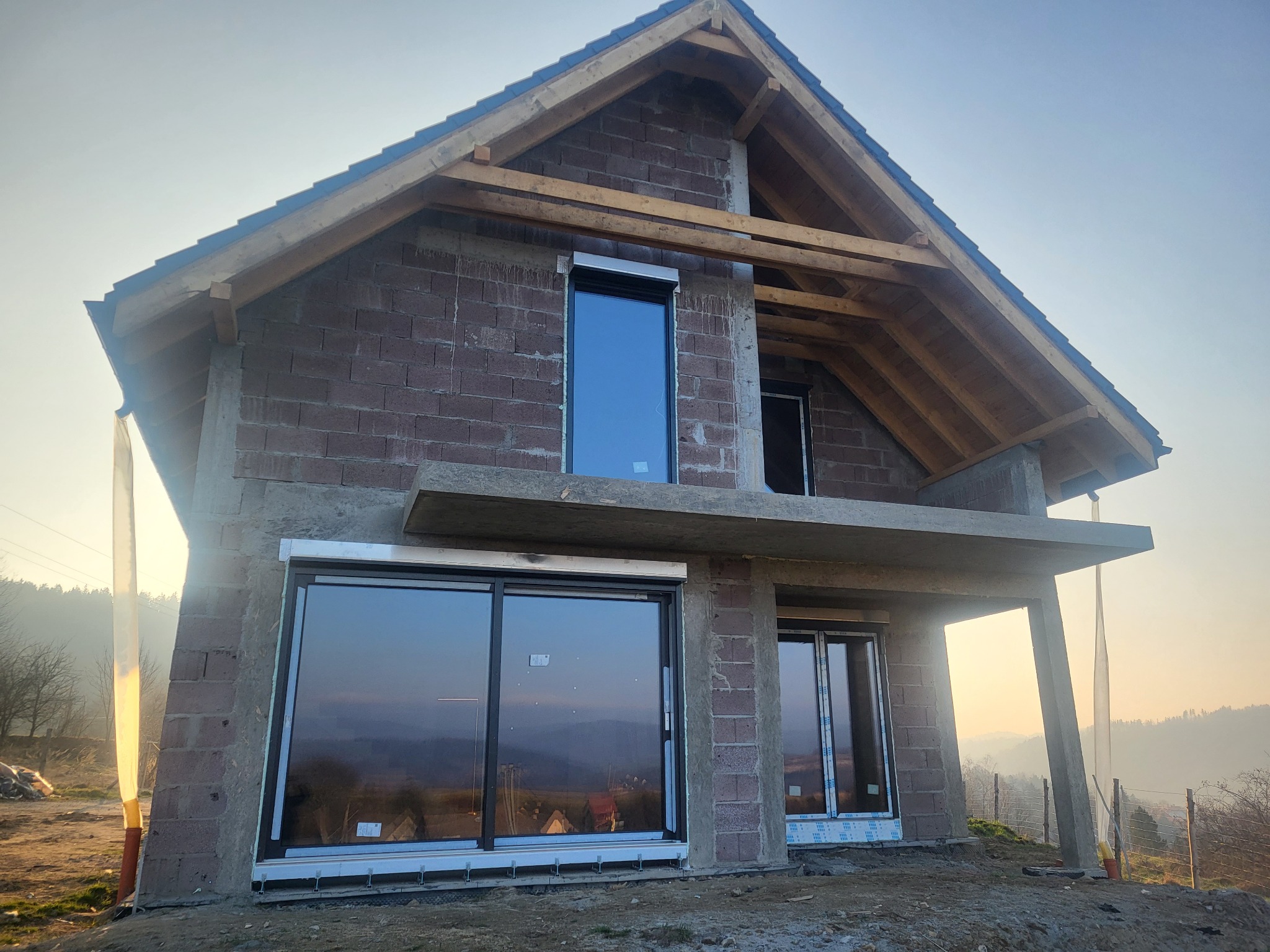Surowy stan domu jednorodzinnego z keramzytobetonu, widoczne wstawione okna, niezakończona konstrukcja dachu i elementy szalunków.