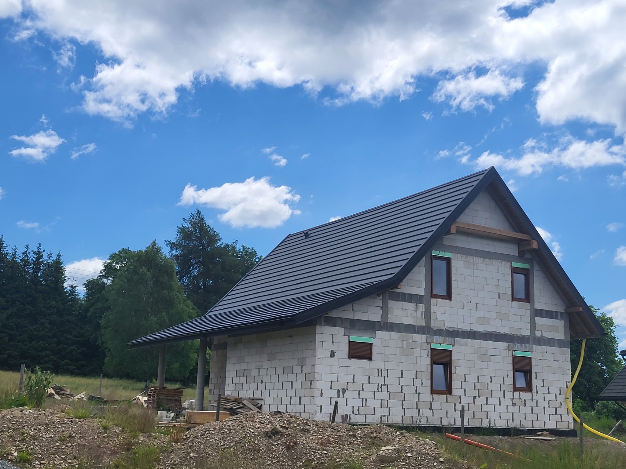 Surowy, dwukondygnacyjny budynek z keramzytobetonowych bloczków, z ciemną dachówką, w trakcie budowy, z widocznymi drewnianymi elementami konstrukcyjnymi i oknami z roletami, na tle błękitnego...