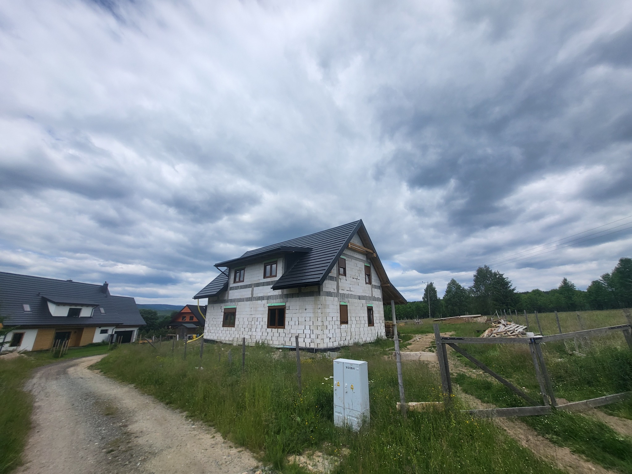 Surowy stan dwukondygnacyjnego domu z białych bloczków, z ciemnym dachem i brązowymi oknami, na tle pochmurnego nieba, z widoczną skrzynką elektryczną i polną drogą.