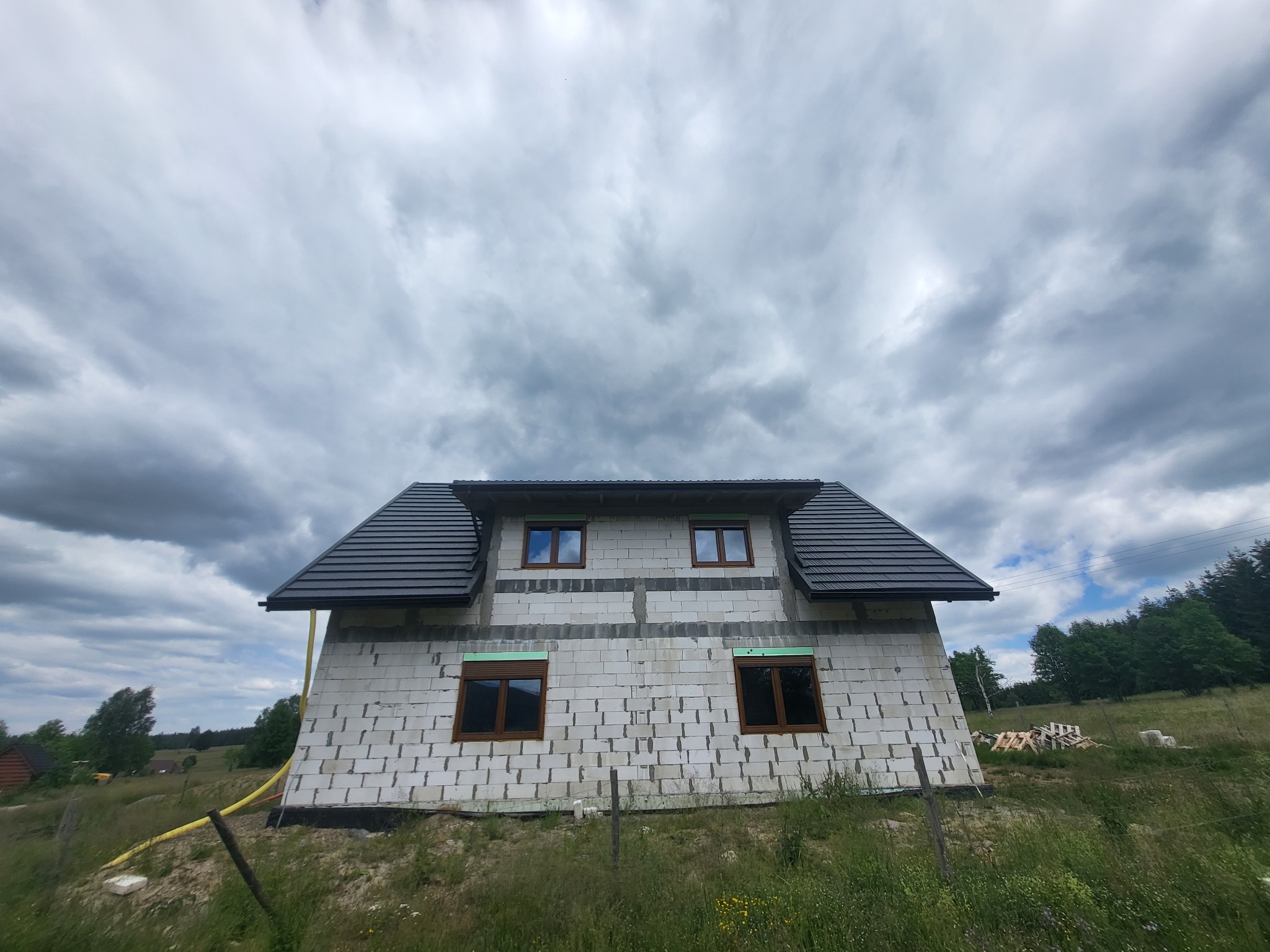 Surowy stan dwukondygnacyjnego domu z białych bloczków, z zamontowaną ciemną dachówką i brązowymi oknami, na tle pochmurnego nieba i zielonego terenu.