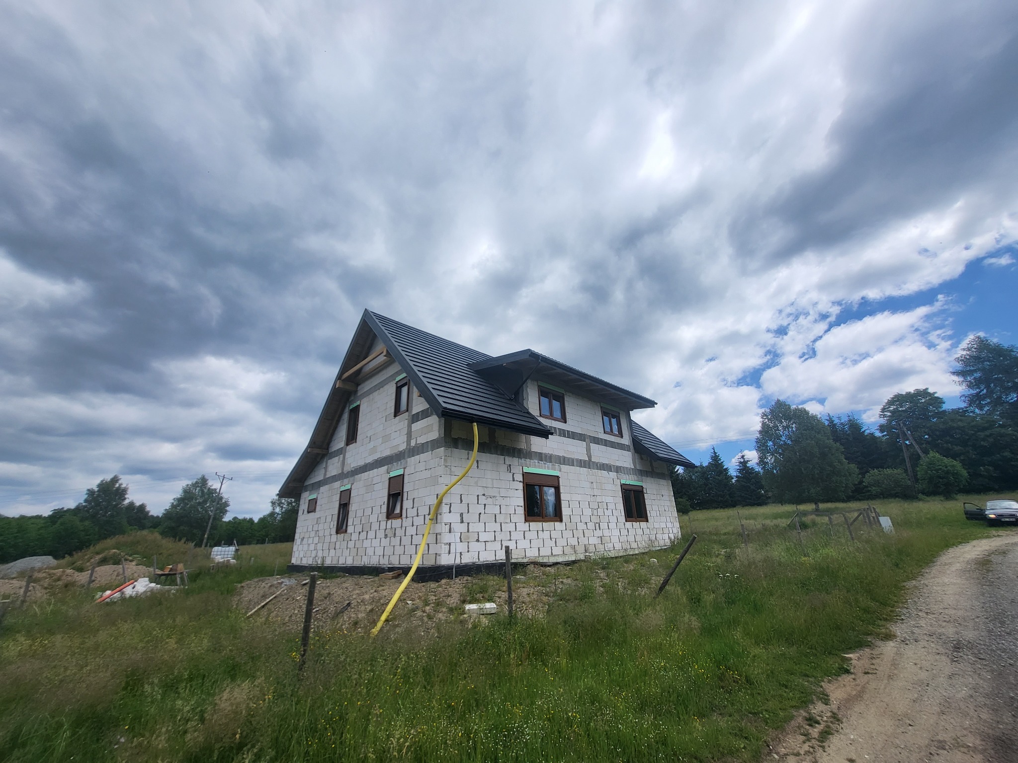 Surowy dom murowany z keramzytobetonu w trakcie budowy, z widoczną konstrukcją dachu, oknami z roletami zewnętrznymi i żółtym wężem na elewacji, na tle pochmurnego nieba i zarośniętej działki.