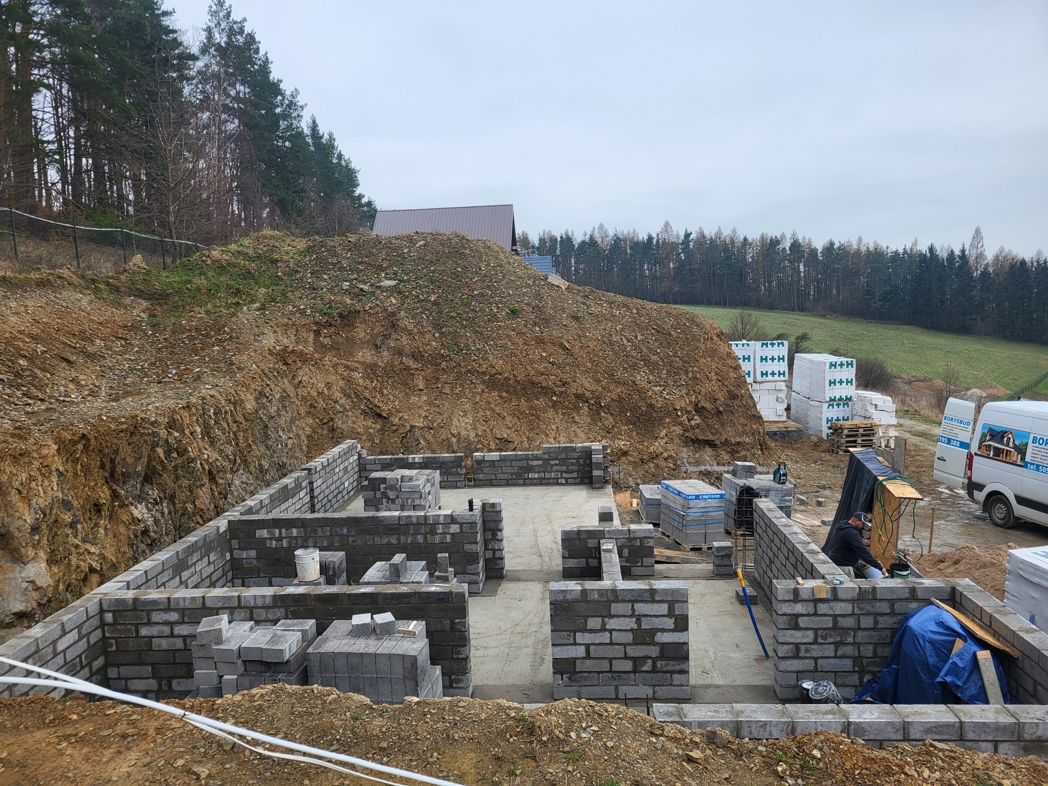 Wznoszenie fundamentów z bloczków betonowych na placu budowy; widoczny pracownik w trakcie wykonywania prac murarskich, zaplecze budowy, hałda ziemi i las w tle.
