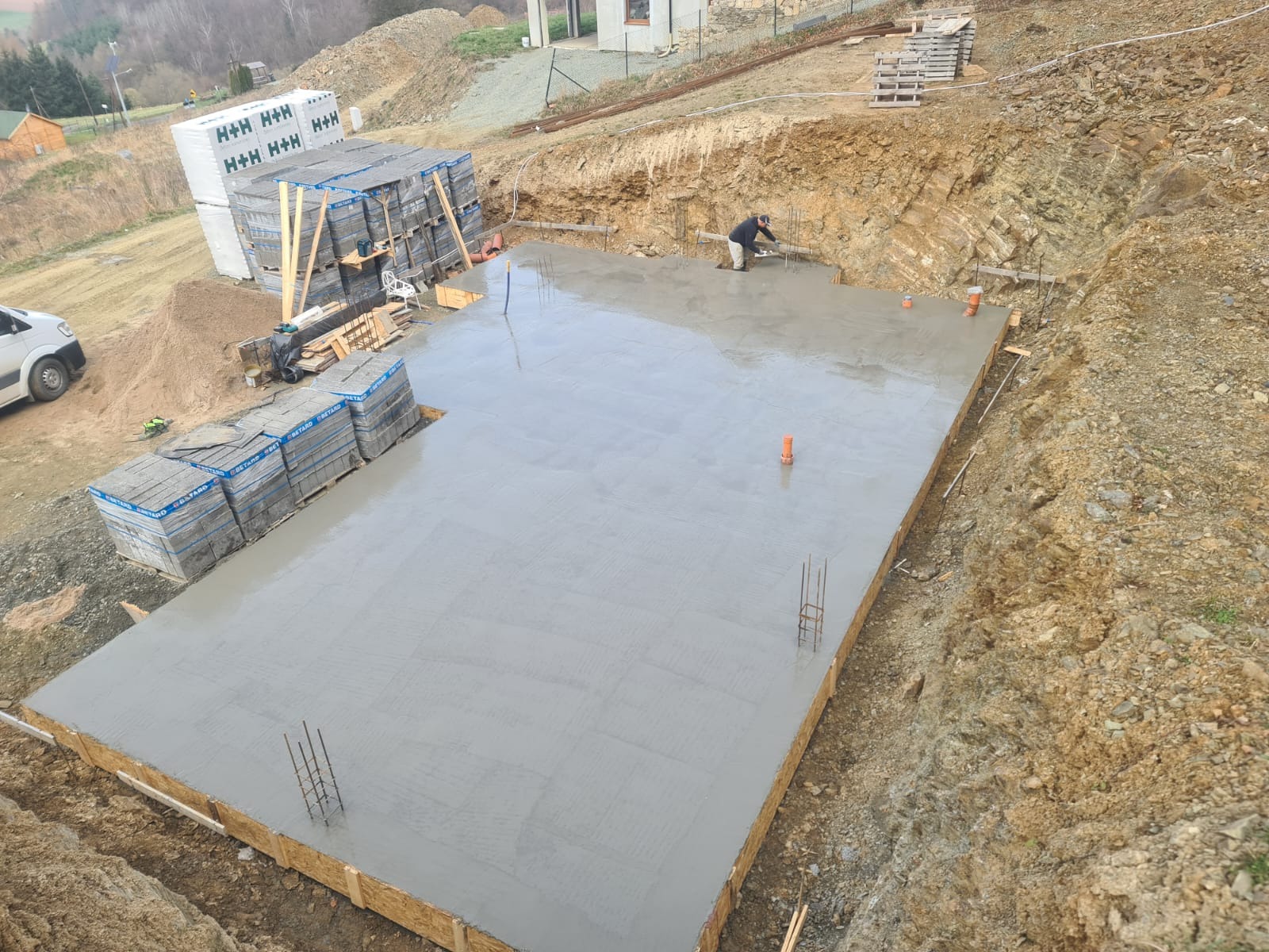 Wylany, świeży beton na przygotowanym fundamencie z widocznymi wystającymi prętami zbrojeniowymi, drewnianym szalunkiem i stosem bloczków budowlanych w tle, podczas prac budowlanych.