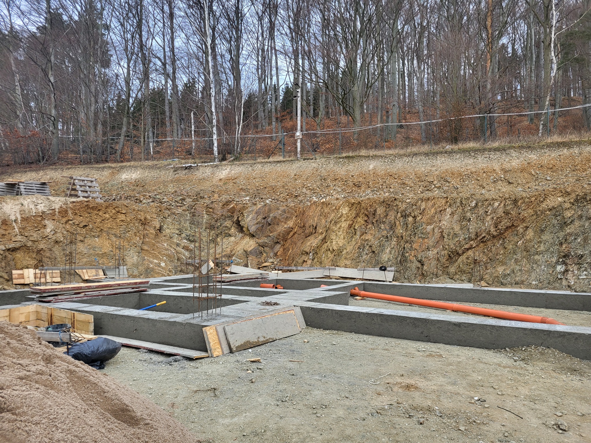 Wylane betonowe ławy fundamentowe z wystającymi prętami zbrojeniowymi, pomarańczowe rury kanalizacyjne wewnątrz, widok na wykop ze skalistym zboczem i lasem bezlistnym w tle.