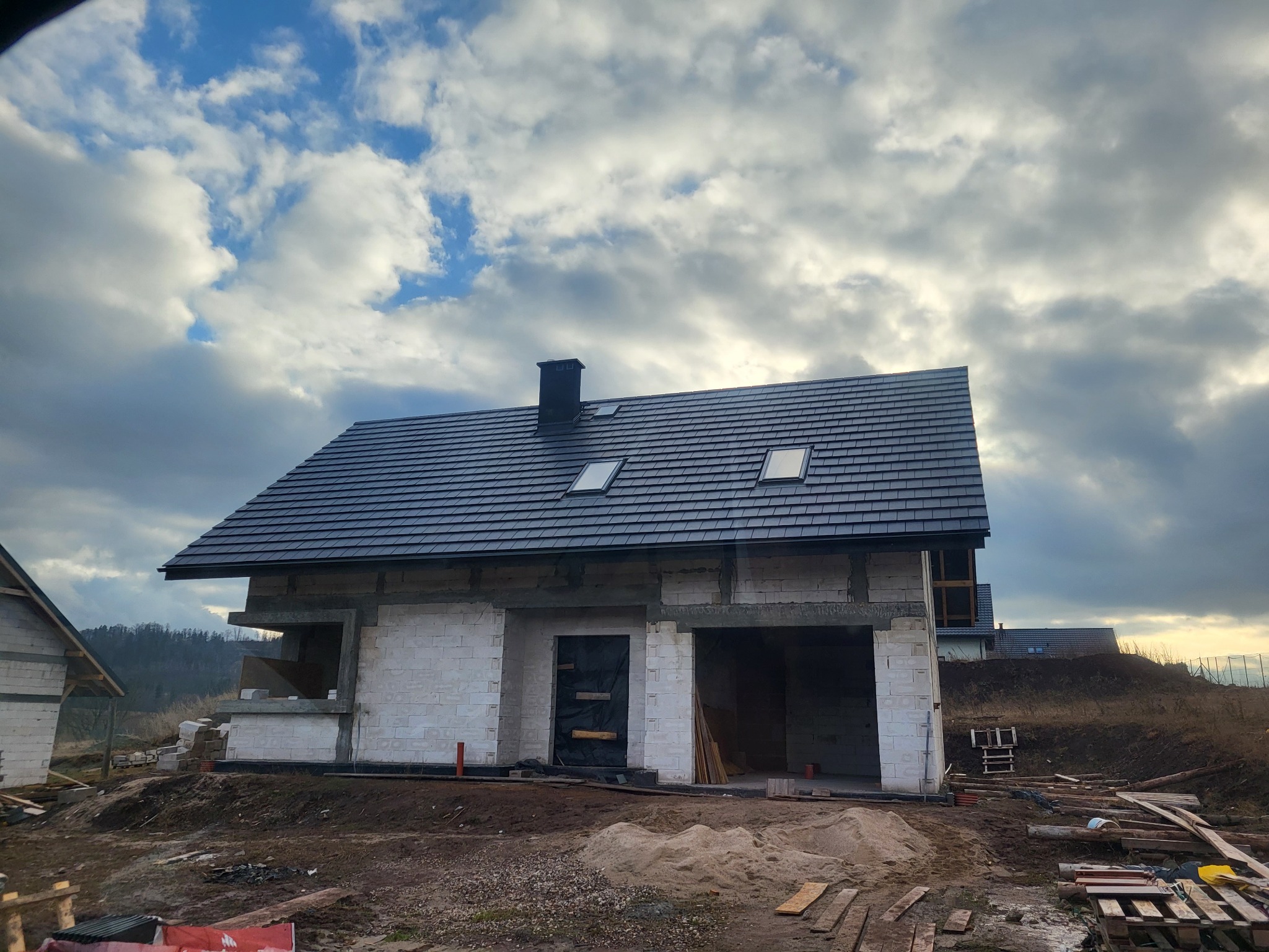 Niedokończony dom z białych bloczków z ciemną dachówką i dwoma oknami dachowymi, widok na budowę w pochmurny dzień.