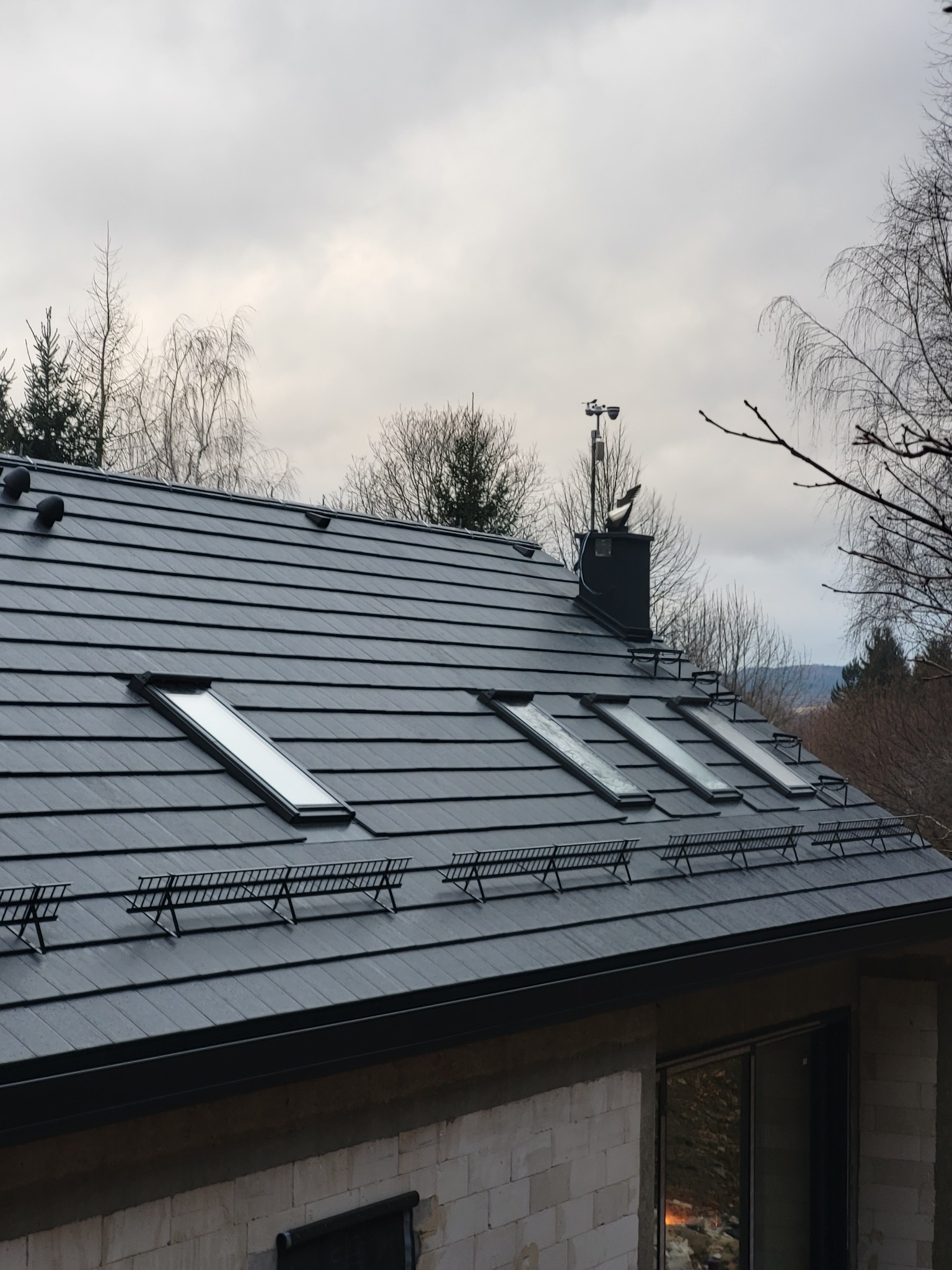 Szary dach z oknami dachowymi i czarnym kominem zwieńczonym figurką orła, widoczny fragment ściany z pustaków i okno. W tle drzewa i pochmurne niebo.