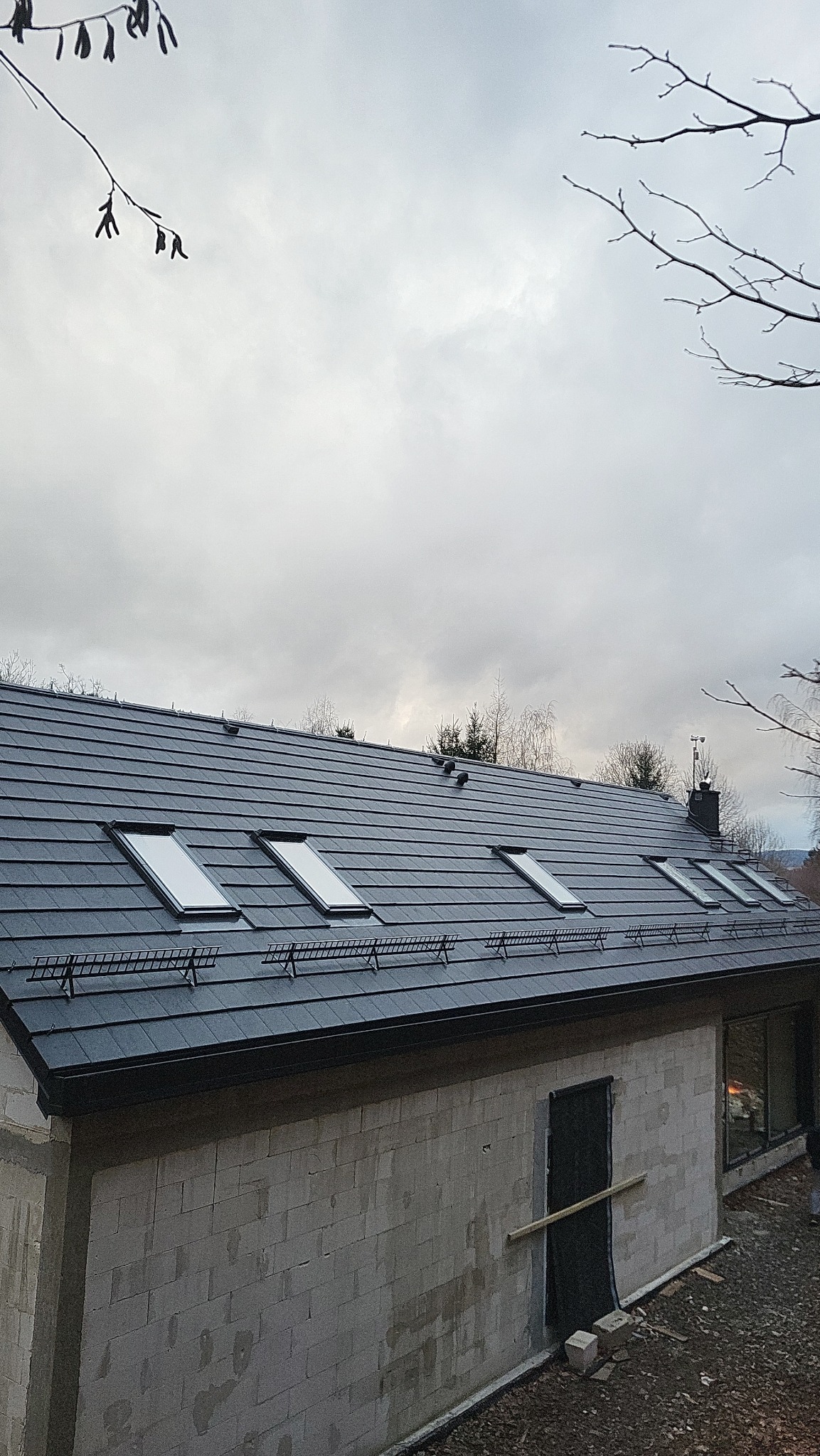 Niedokończony dom z keramzytu z ciemną dachówką i oknami dachowymi, widoczny fragment ściany z otworem na drzwi, pochmurne niebo w tle.