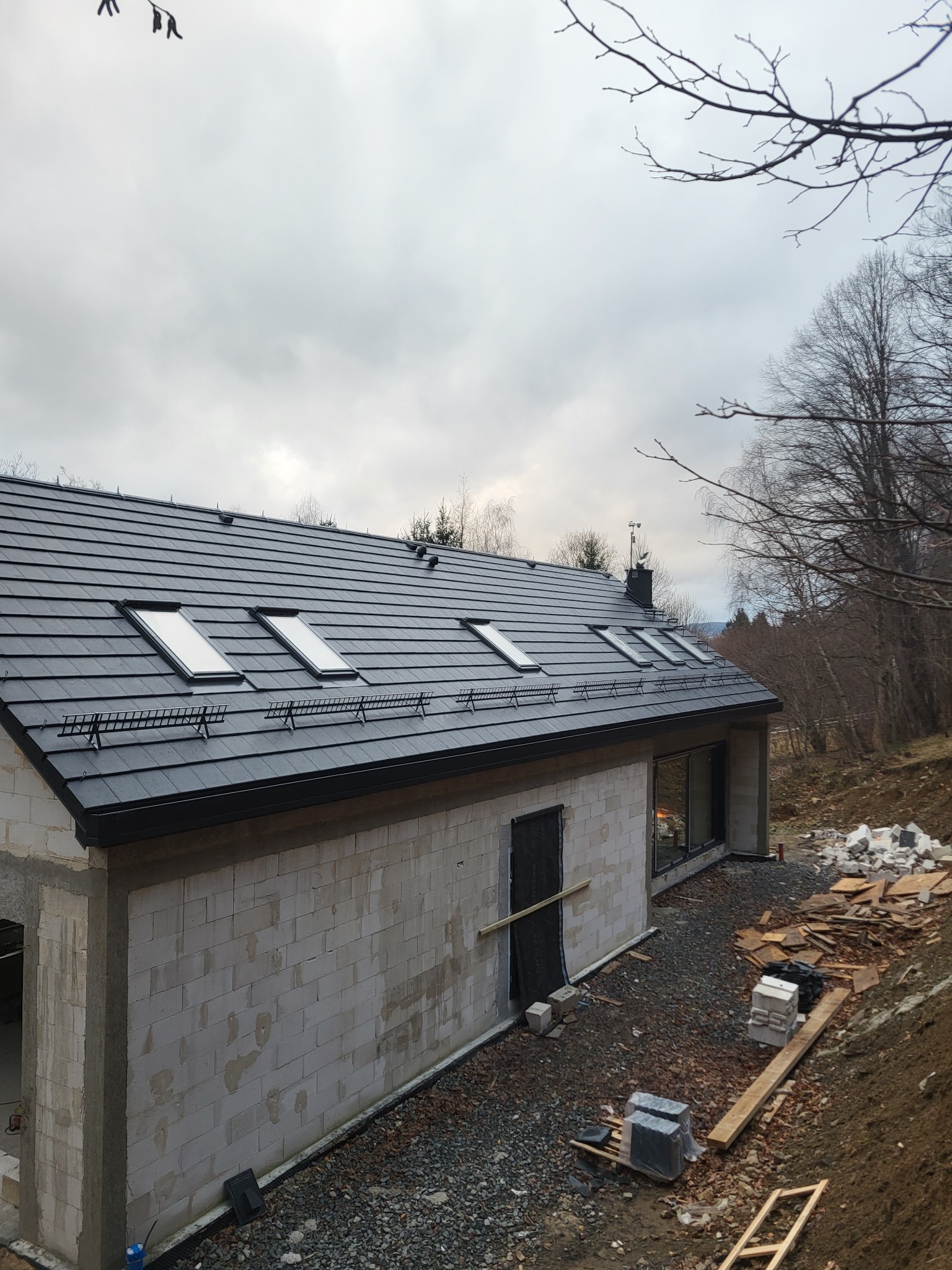 Surowy dom z keramzytu w trakcie budowy, widok z góry. Ciemna dachówka z oknami dachowymi, ściany z pustaków, teren wokół domu przygotowany pod dalsze prace.